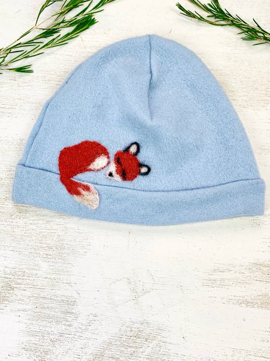 The Sleeping Fox Cashmere Hat - Child by Ash & Rose is a light blue, handmade kids hat crafted from upcycled materials, featuring an embroidered red fox curled up on the front. Shown on a white wooden surface with green sprigs nearby.