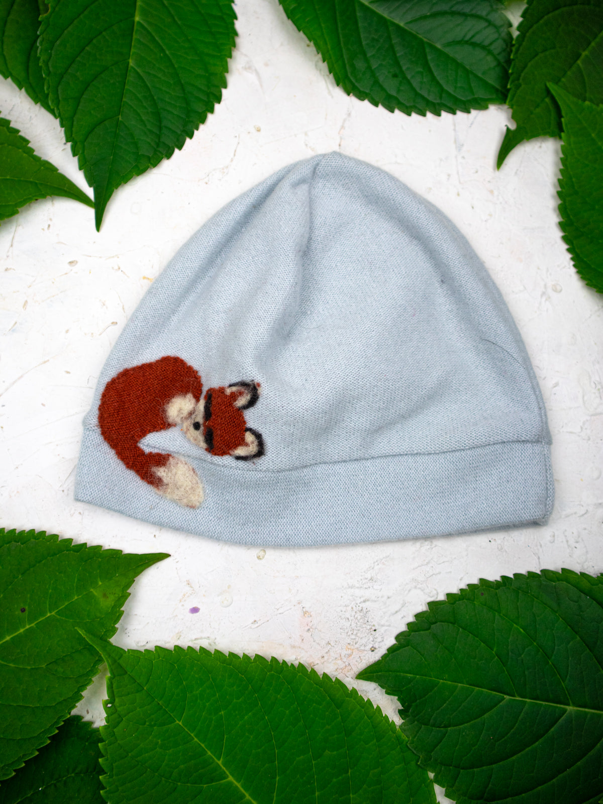 The Ash & Rose Sleeping Fox Cashmere Hat - Child, in light blue with an embroidered sleeping fox, is displayed on a white surface surrounded by large green leaves.