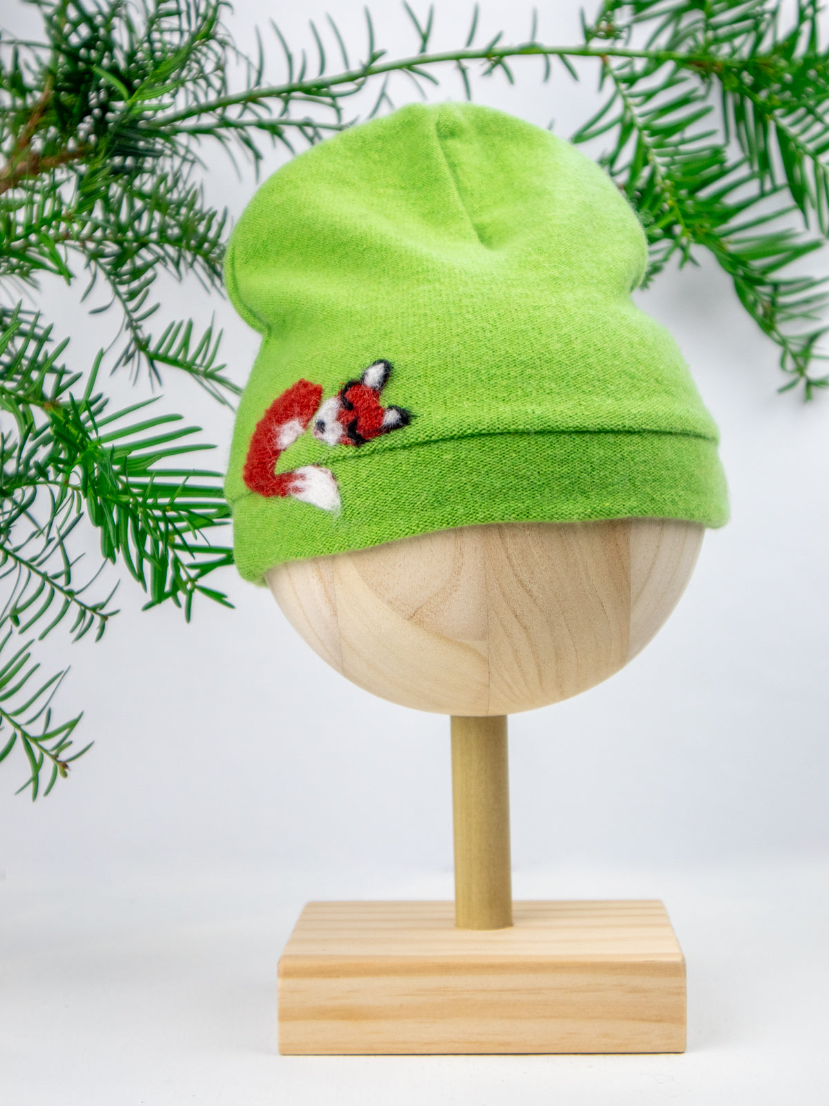 The Ash & Rose Sleeping Fox Cashmere Hat - Child is a lime green, handmade beanie from upcycled materials with an embroidered orange and white fox, shown on a wooden mannequin head with pine branches in the background.