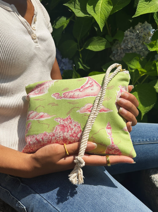 Wearing a white ribbed shirt and jeans, a person holds the Ash & Rose Nantucket Purse—a limited edition green pouch with pink patterns and a thick white rope handle—while sitting outdoors among leafy plants.