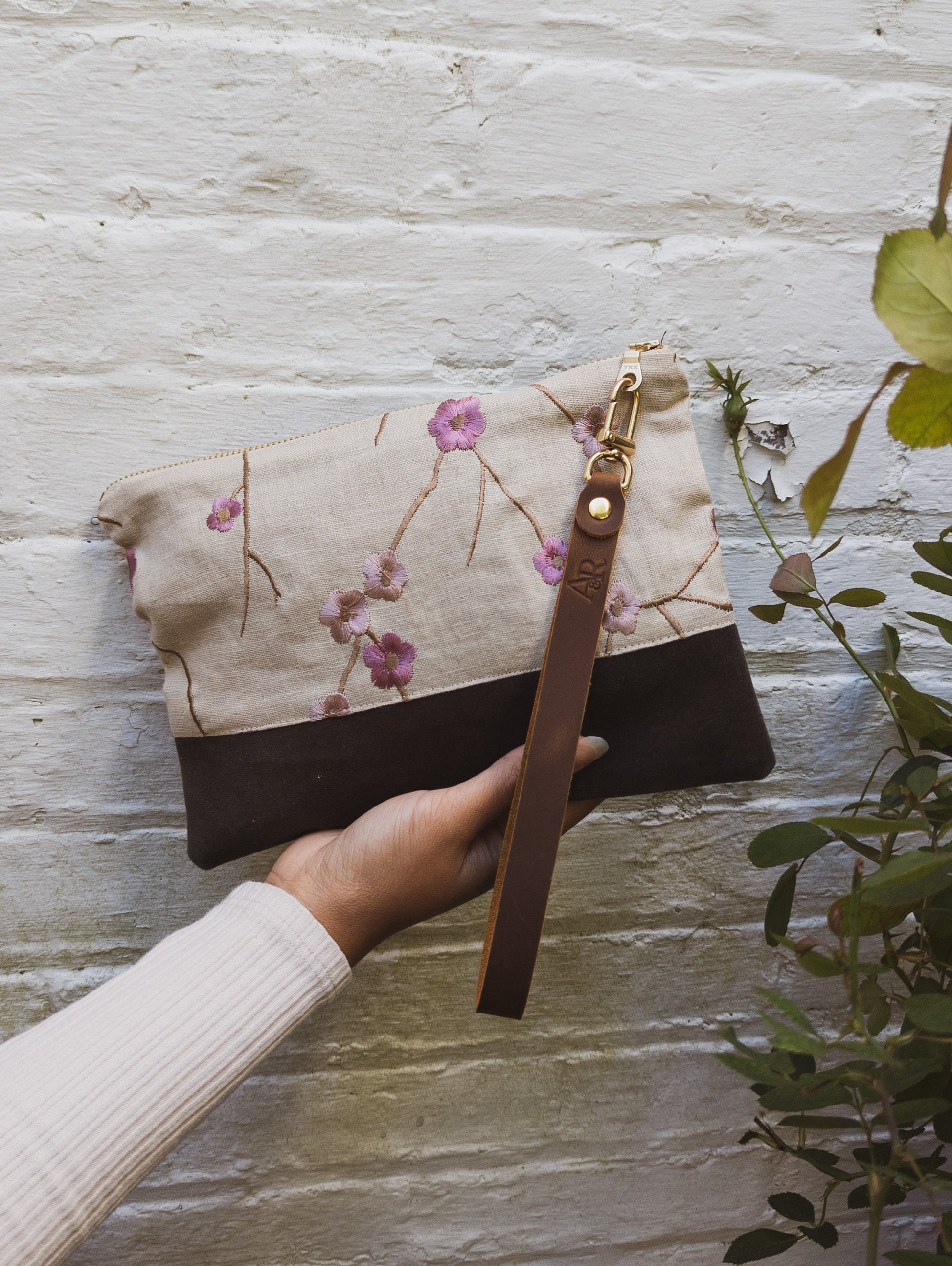 A hand holds the limited edition Alice Embroidered Linen Wristlet by Ash & Rose, featuring vintage linen with floral embroidery and a brown leather strap. The backdrop is a white brick wall with green leafy plants nearby.