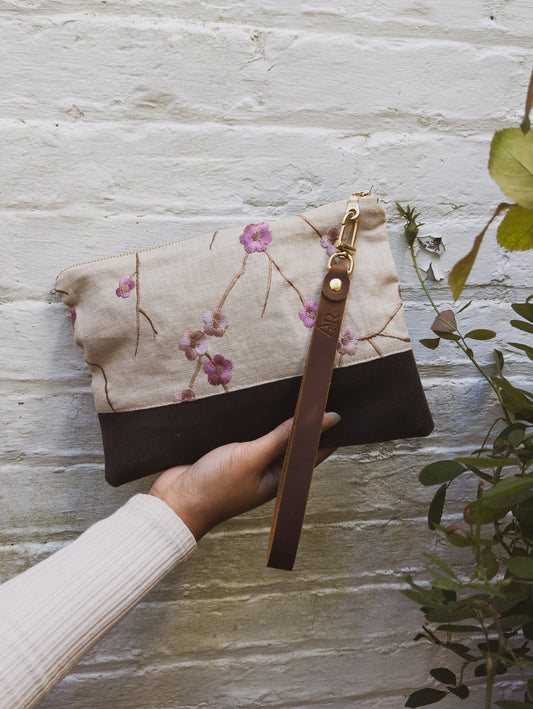A hand holds the limited edition Alice Embroidered Linen Wristlet by Ash & Rose, featuring vintage linen with floral embroidery and a brown leather strap. The backdrop is a white brick wall with green leafy plants nearby.