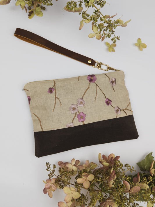 The Alice Embroidered Linen Wristlet by Ash & Rose features beige and dark brown floral embroidery, a leather strap, and vintage linen, displayed with dried hydrangea on a white background.