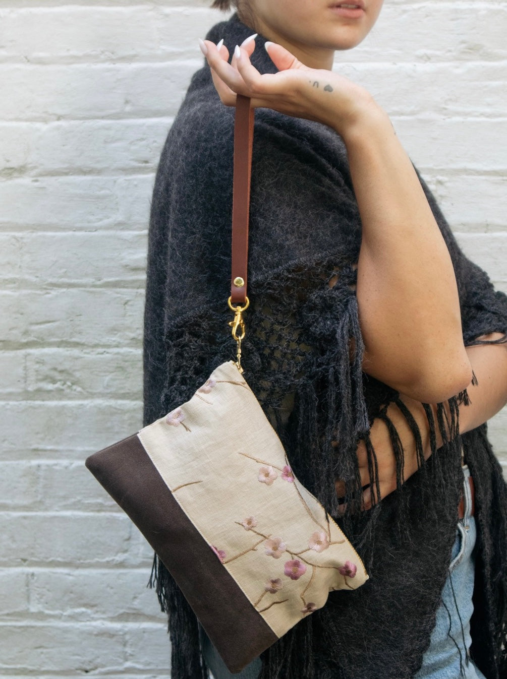 A person in a dark fringed shawl holds the Ash & Rose Alice Embroidered Linen Wristlet, featuring a brown strap and beige accents, against a light brick wall. Only the lower half of their face is visible.