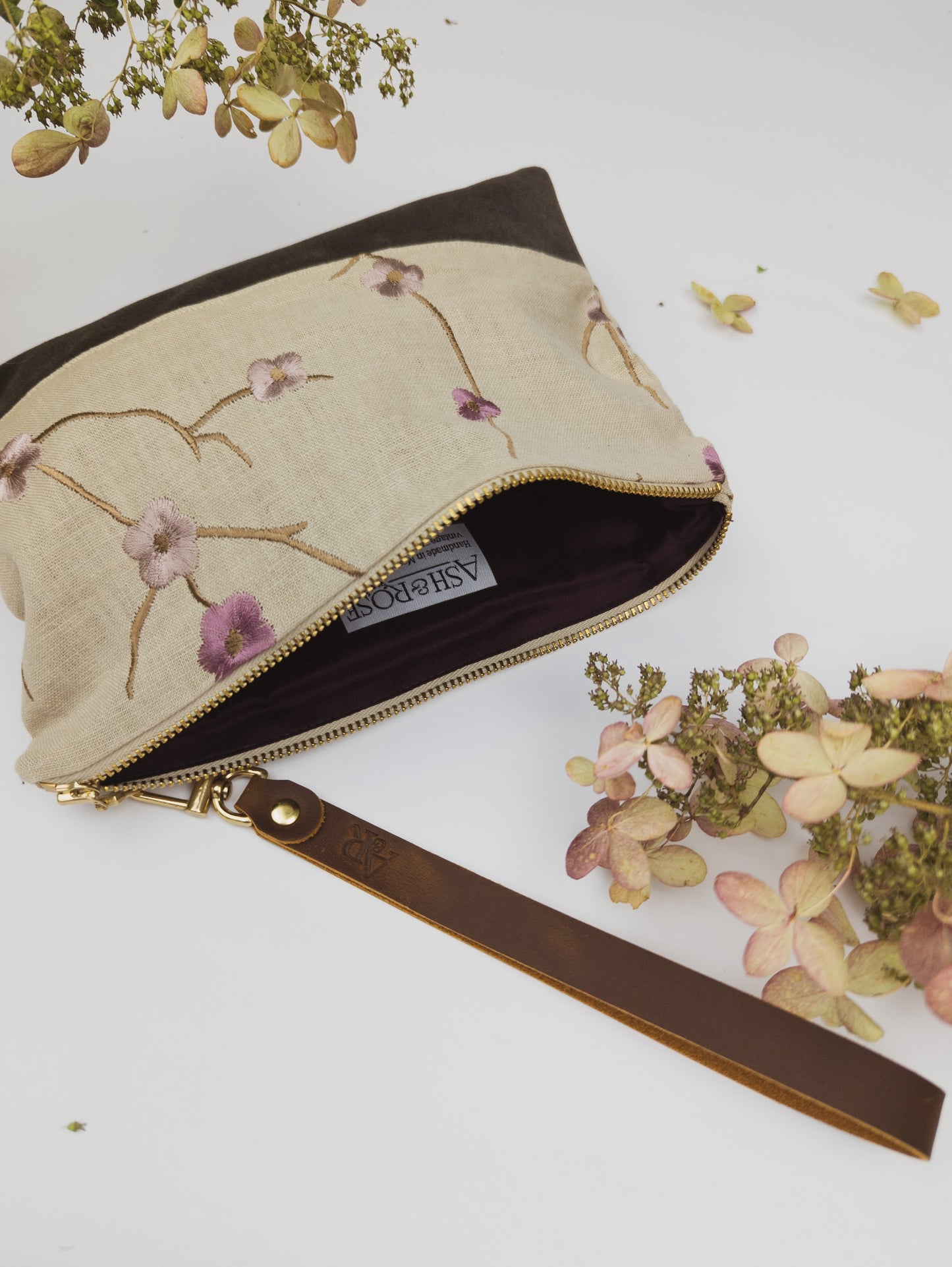 The Alice Embroidered Linen Wristlet by Ash & Rose is a limited edition fabric clutch with purple and pink floral embroidery on vintage linen, a brown leather wrist strap, and a gold zipper, shown partly open amid dried flowers on white.