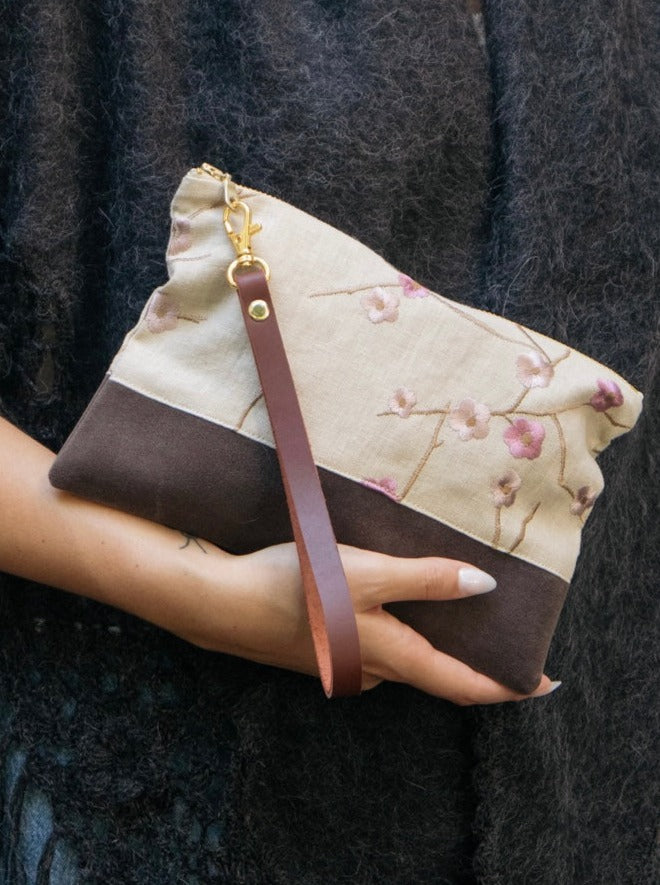 A person holds the Alice Embroidered Linen Wristlet by Ash & Rose, a beige and brown floral accessory with a brown leather wrist strap, crafted from vintage linen, set against a textured dark background.