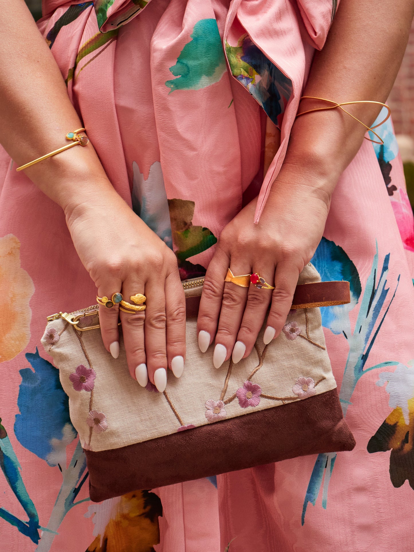 A woman in a pink floral dress holds the limited-edition Alice Embroidered Linen Wristlet by Ash & Rose. Her long white nails, colorful rings, and gold bracelets complete her stylish look.