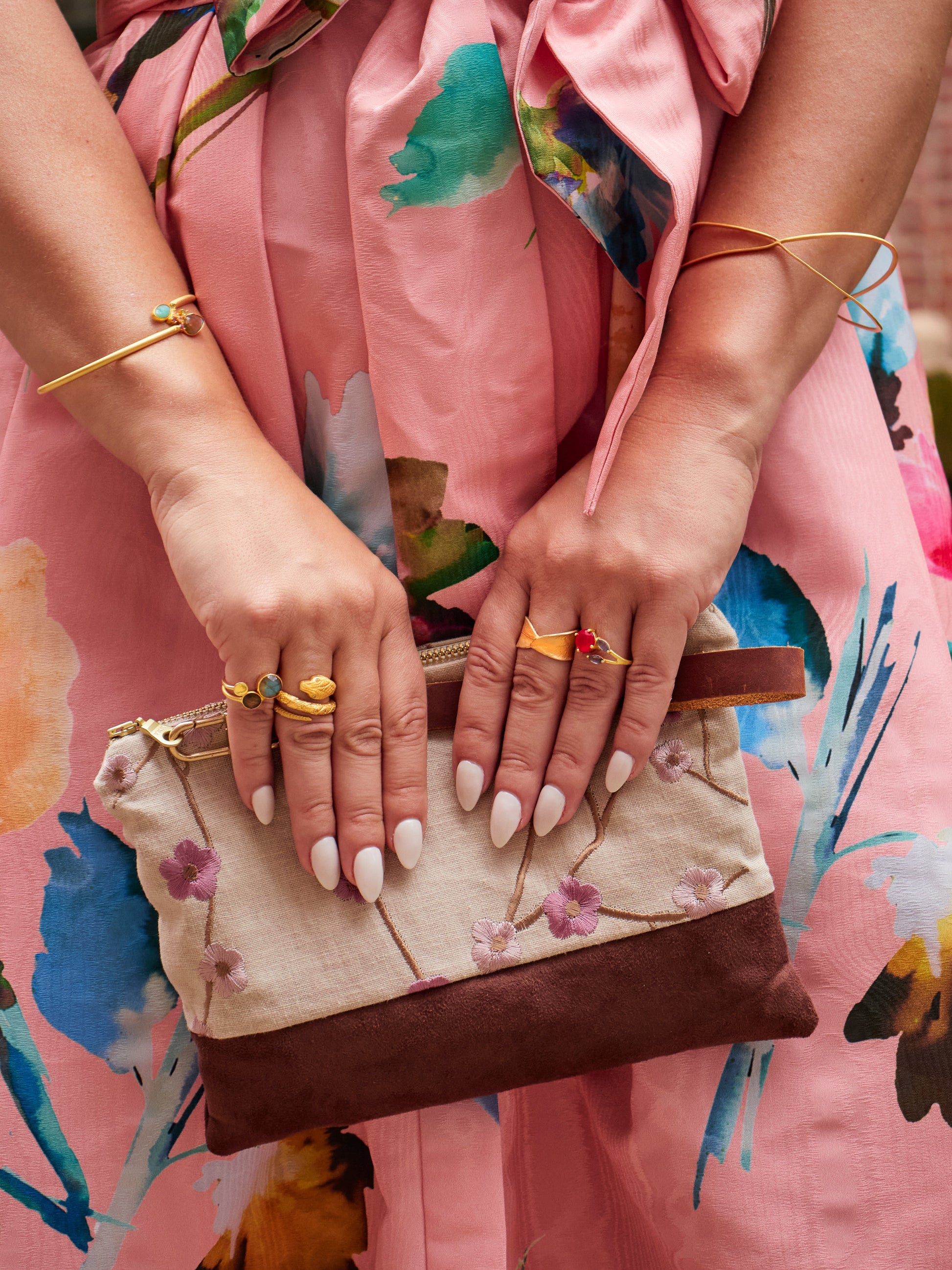 A woman in a pink floral dress holds the limited-edition Alice Embroidered Linen Wristlet by Ash & Rose. Her long white nails, colorful rings, and gold bracelets complete her stylish look.