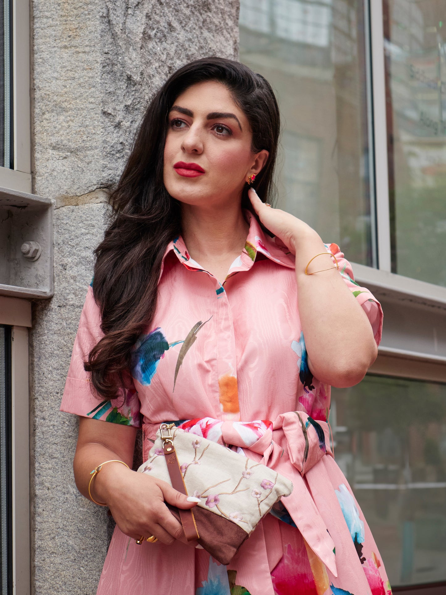 A woman with long dark hair wears a pink floral dress and holds the Ash & Rose Alice Embroidered Linen Wristlet as she stands by a stone wall and glass window, gazing thoughtfully to the side with one hand touching her neck.