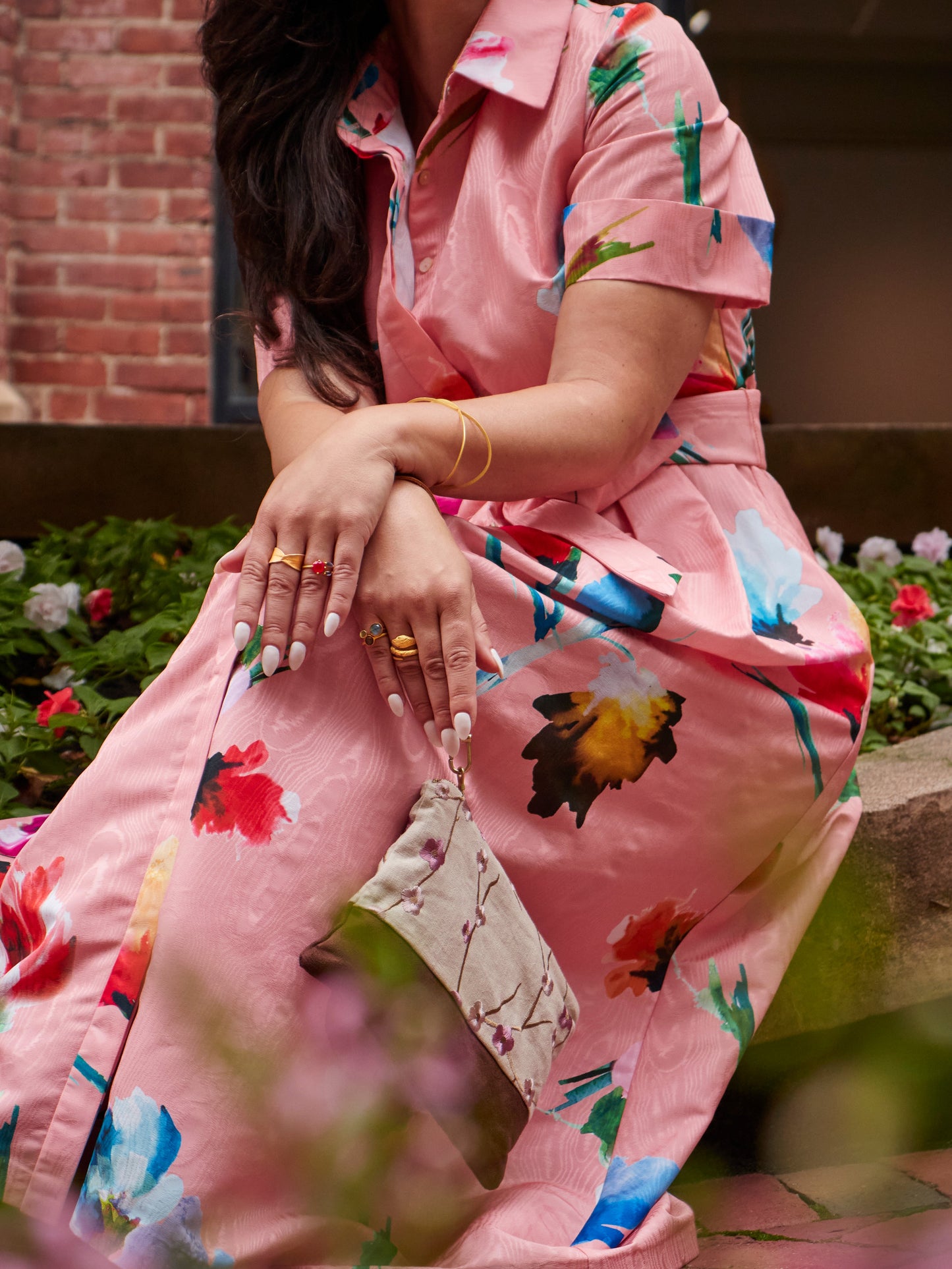 A woman in a pink floral dress sits outdoors, surrounded by colorful flowers and brick walls. She holds the Ash & Rose Alice Embroidered Linen Wristlet, wears gold jewelry, and her manicured nails show as her hair partially covers her face.