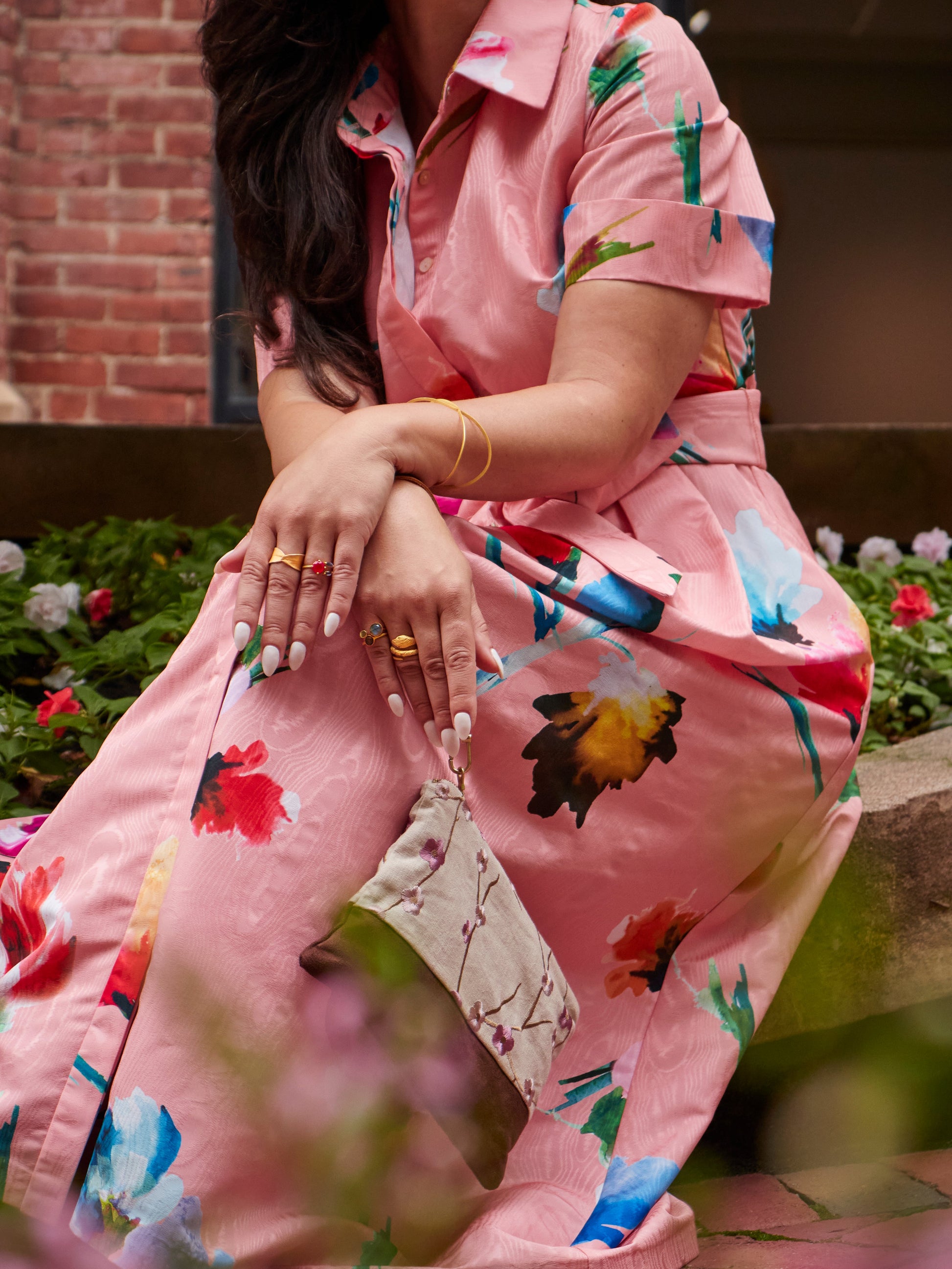 A woman in a pink floral dress sits outdoors, surrounded by colorful flowers and brick walls. She holds the Ash & Rose Alice Embroidered Linen Wristlet, wears gold jewelry, and her manicured nails show as her hair partially covers her face.