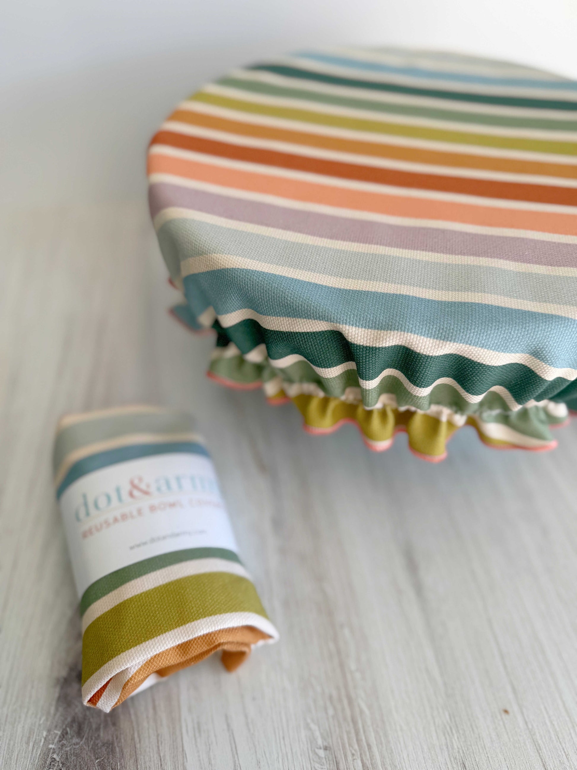 A Dot and Army Reusable Bowl Cover in Beach Chair Stripes is stretched over a bowl, with a matching folded bread proofing cover beside it on a light wooden surface.