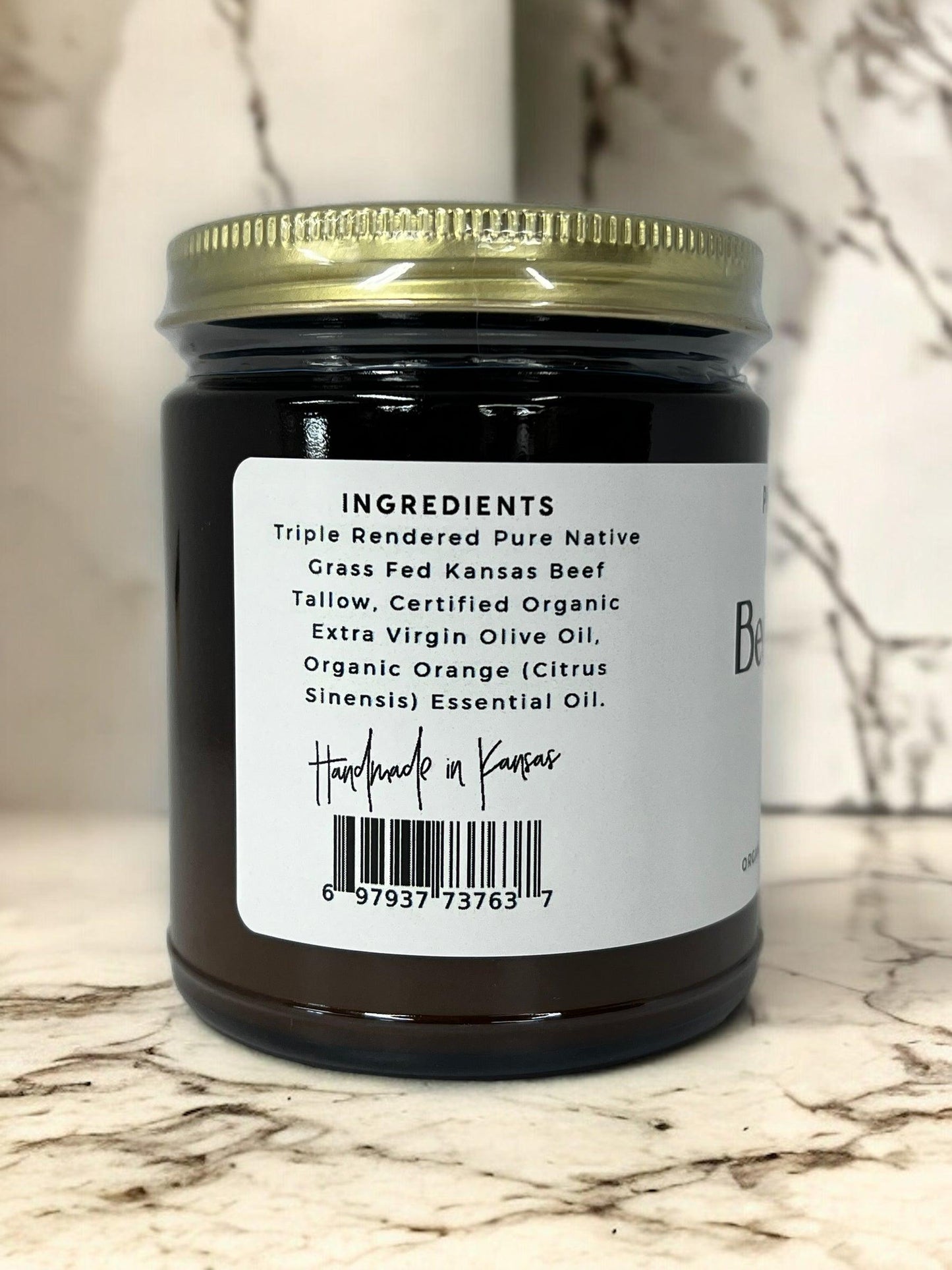 A dark glass jar with a gold lid from Prairie Fire Tallow, Candles, and Lavender sits on marble. The label for Beef Tallow Balm lists organic grass-fed tallow, olive oil, and orange oil, and reads “Handmade in Kansas.”.
