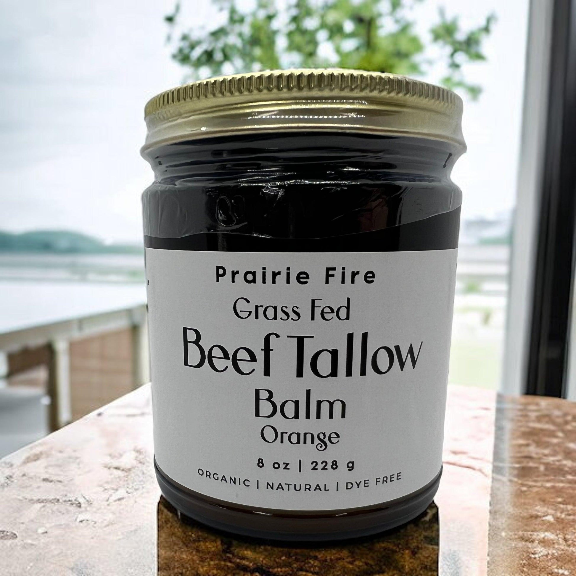 A jar of Prairie Fire Tallow, Candles, and Lavender Beef Tallow Balm (8 oz) sits on a countertop. Made in the USA from organic grass-fed beef, this natural, dye-free moisturizer is perfect for skin care. Outdoor trees blur through the window behind it.