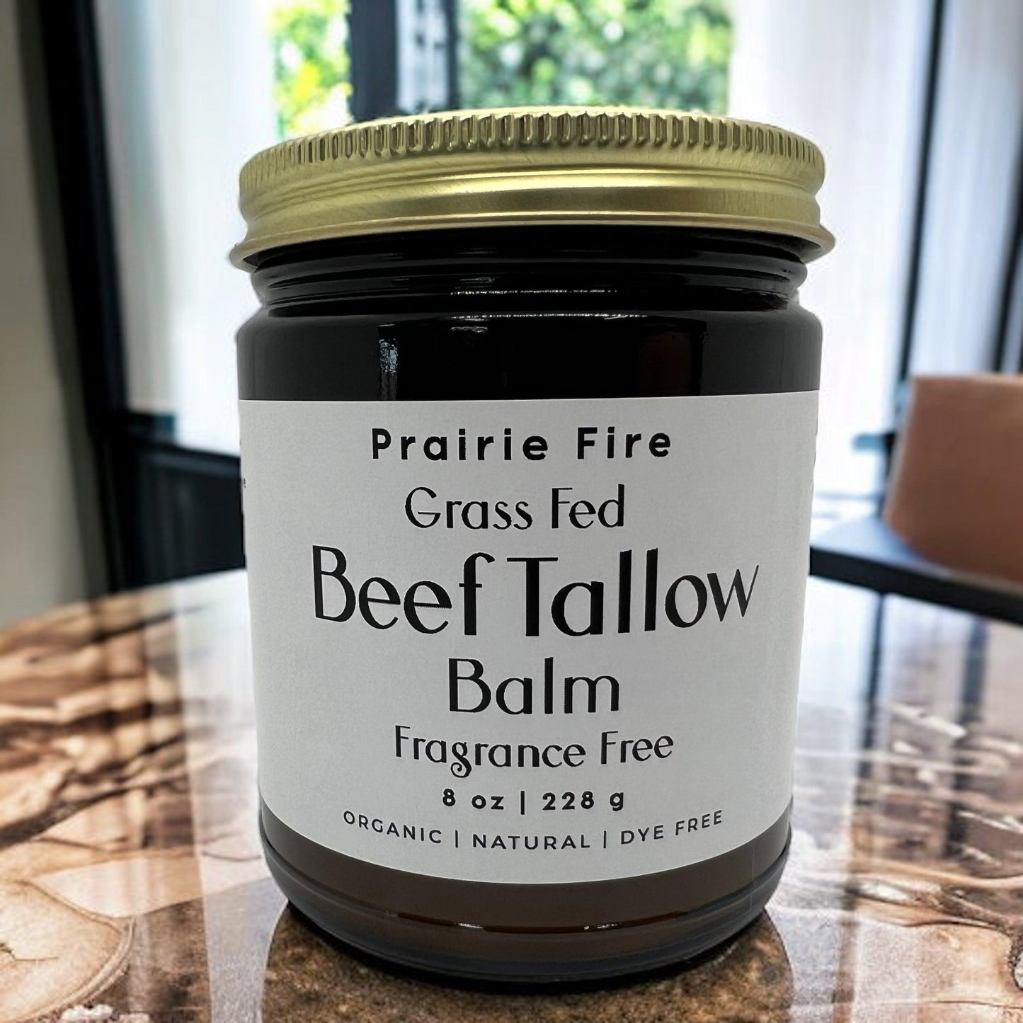 An 8 oz jar of Prairie Fire Tallow, Candles, and Lavender Beef Tallow Balm—organic, grass fed and finished, made in the USA—is shown on a marble surface. The fragrance- and dye-free moisturizer has a window and sofa in the background.