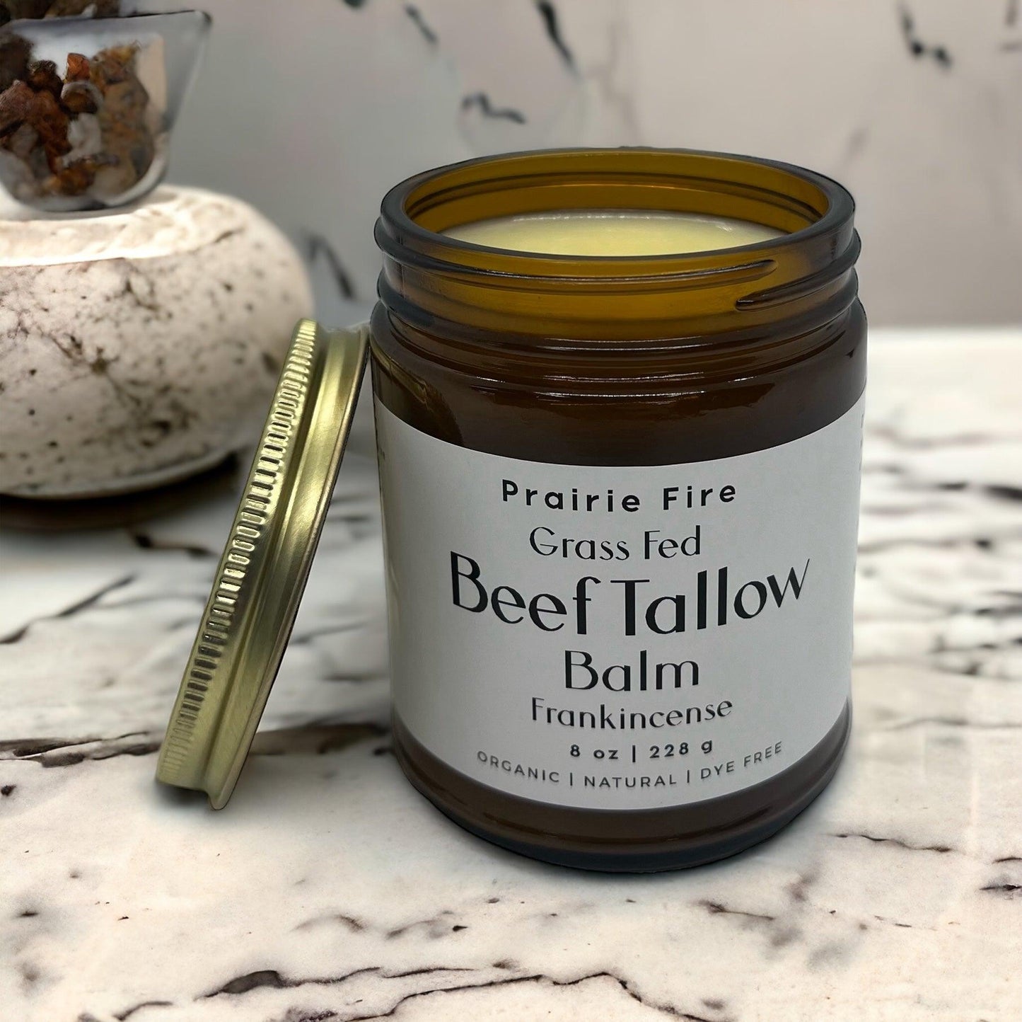 A brown glass jar labeled Prairie Fire Tallow’s Beef Tallow Balm (8 oz), Made in the USA from organic, grass-fed beef tallow, sits on marble with a gold lid, offering natural moisturizing skin care.