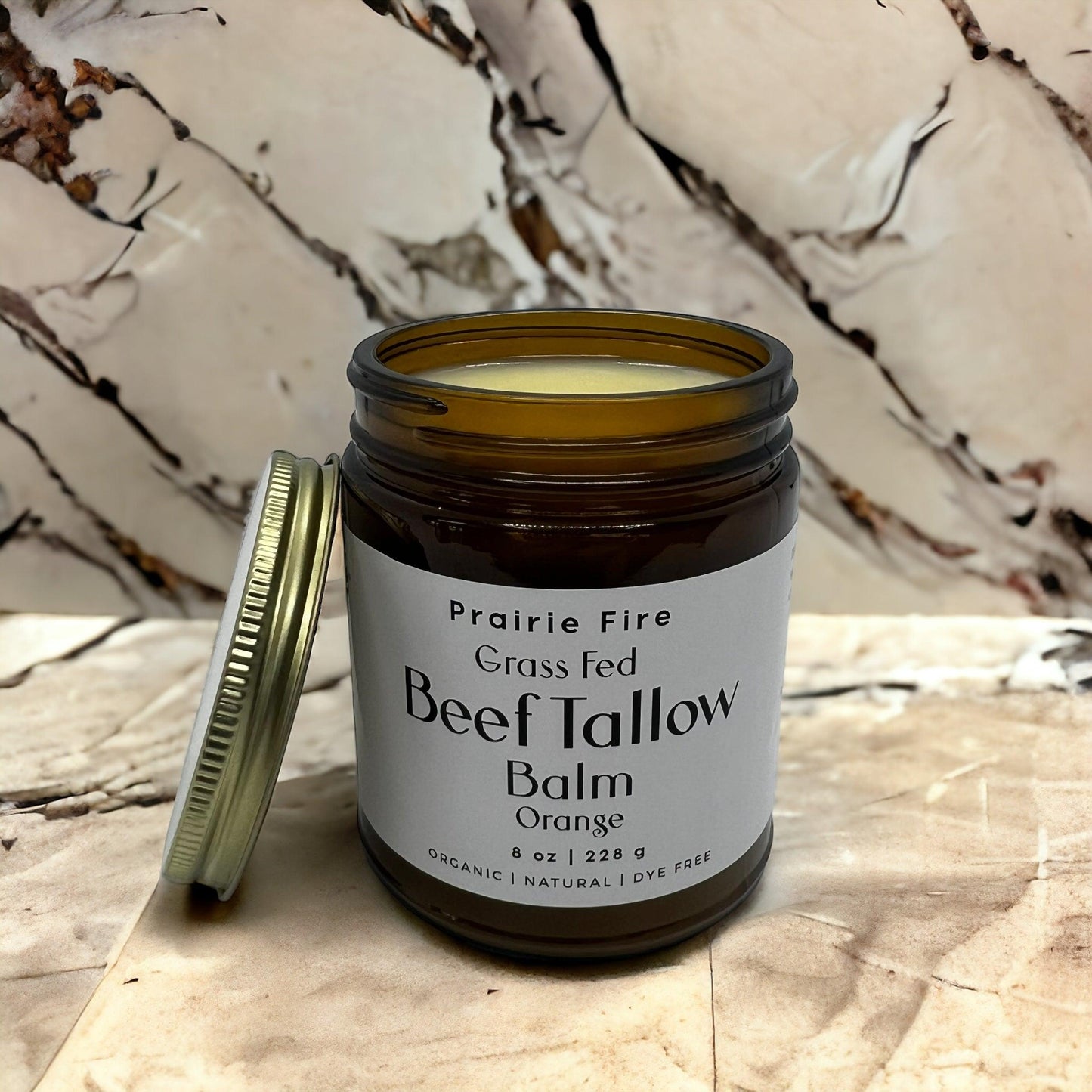 An open jar of Prairie Fire Tallow, Candles, and Lavender Beef Tallow Balm (8 oz) sits on a marble surface with the lid beside it. This organic, grass-fed moisturizer is light yellow and made in the USA—dye-free skin care for nourishing results.
