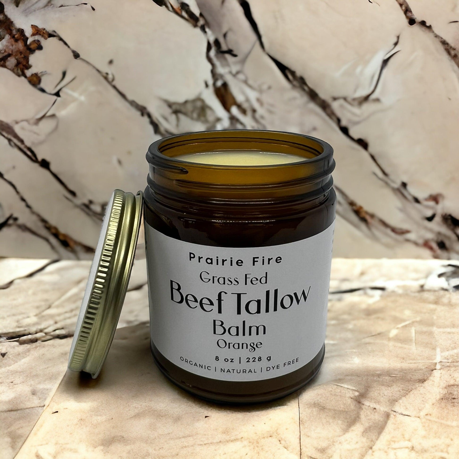 An open jar of Prairie Fire Tallow, Candles, and Lavender Beef Tallow Balm (8 oz) sits on a marble surface with the lid beside it. This organic, grass-fed moisturizer is light yellow and made in the USA—dye-free skin care for nourishing results.