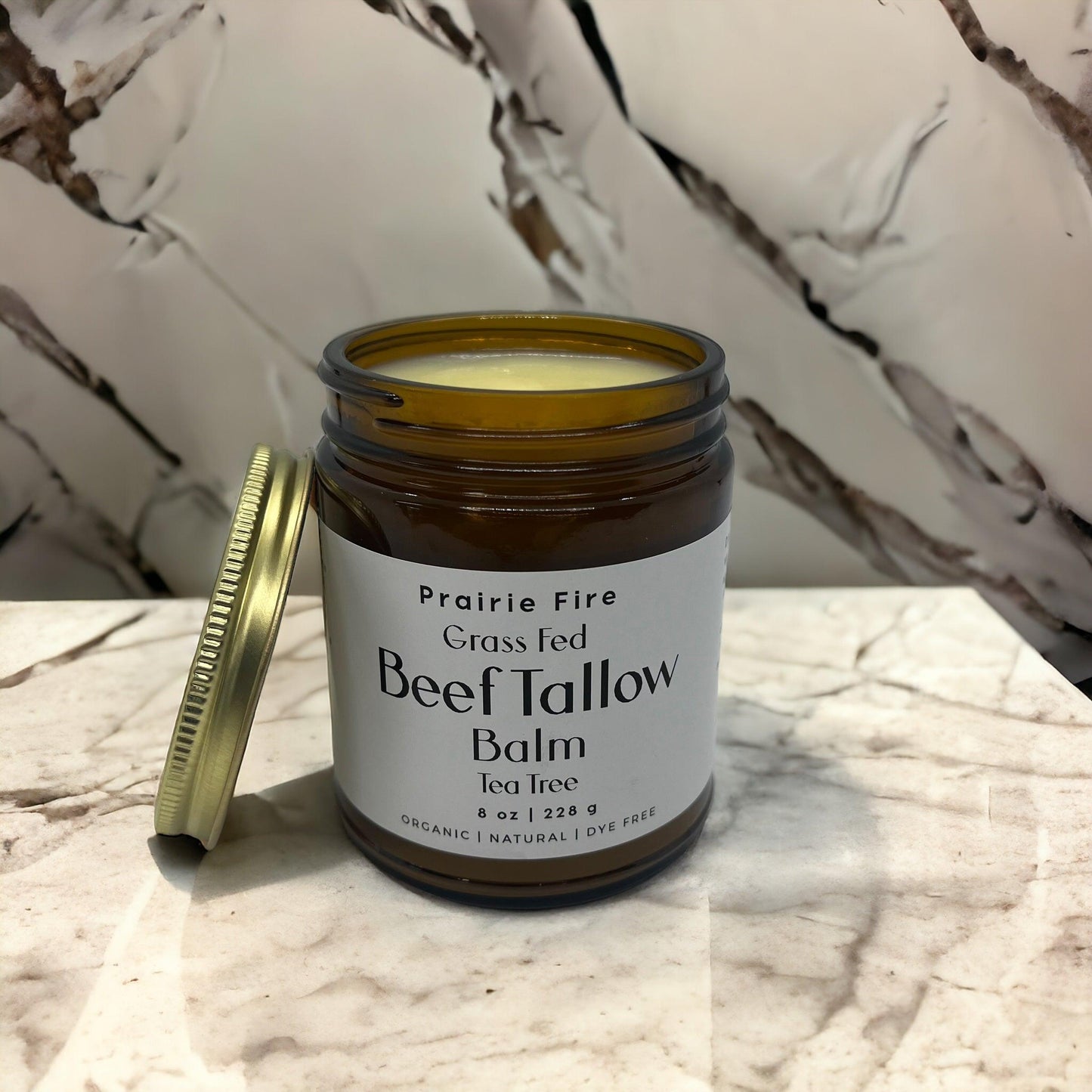 An open jar of Prairie Fire Tallow, Candles, and Lavender Beef Tallow Balm (8 oz, organic grass-fed and finished, made in USA) sits with its gold lid on a marble surface against a marble-patterned background.