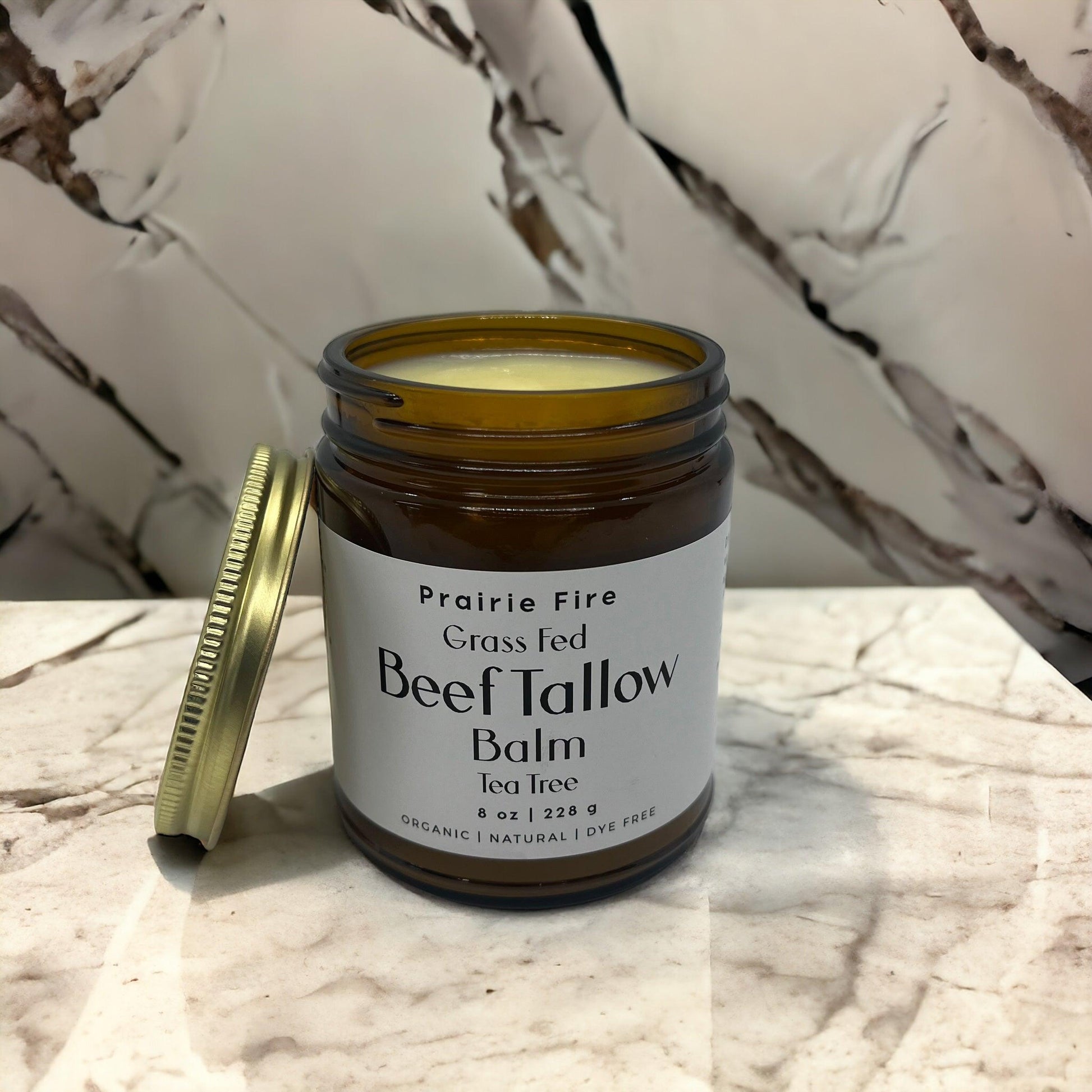 An open jar of Prairie Fire Tallow, Candles, and Lavender Beef Tallow Balm (8 oz, organic grass-fed and finished, made in USA) sits with its gold lid on a marble surface against a marble-patterned background.