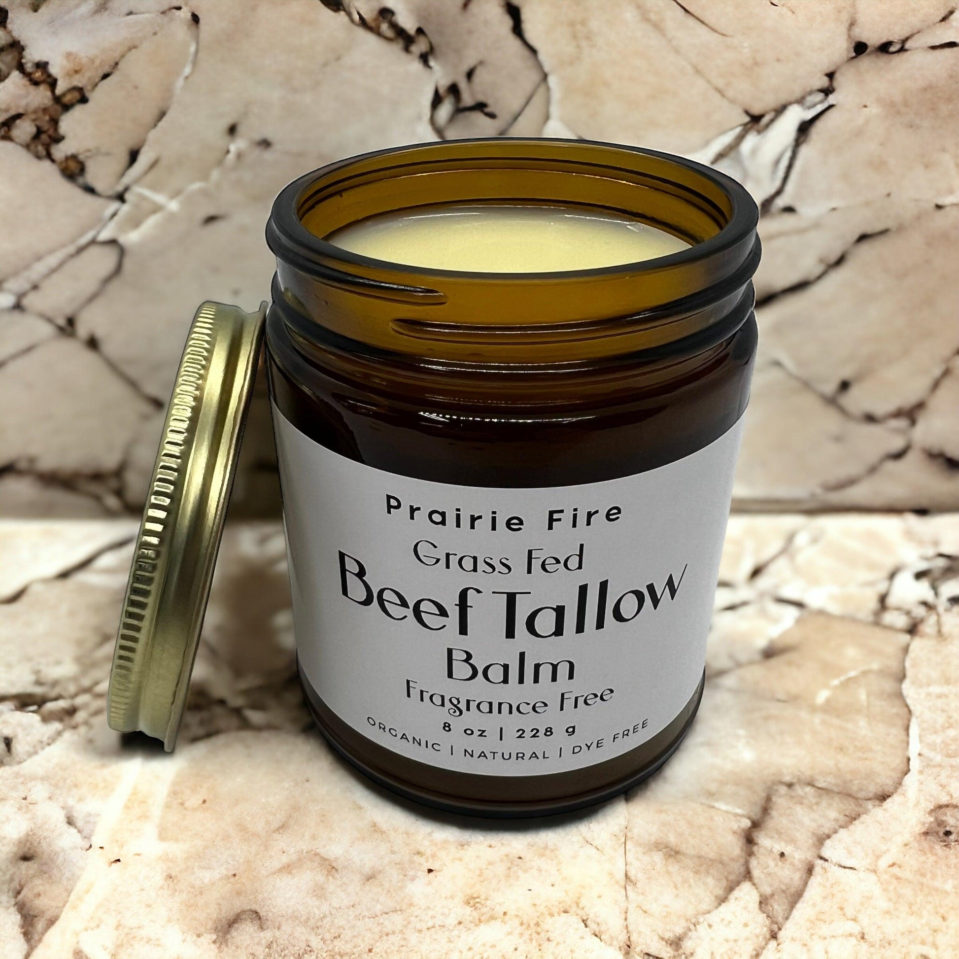An open amber jar of Prairie Fire Tallow, Candles, and Lavender Beef Tallow Balm—8 oz, organic grass fed and finished, made in the USA—sits on marble with its gold lid nearby. The smooth pale balm is a natural skin moisturizer.