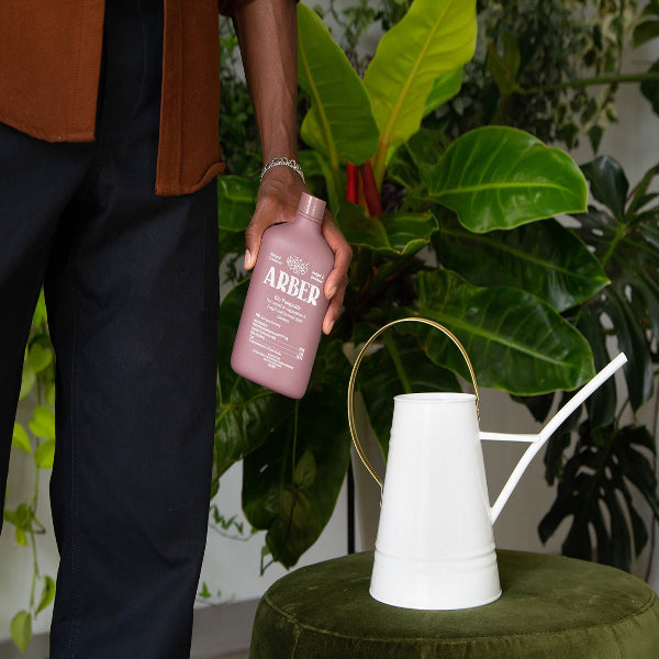 A person holds a bottle of Arber Organic Bio Fungicide beside a white watering can on a green surface, with vibrant houseplants in the background.