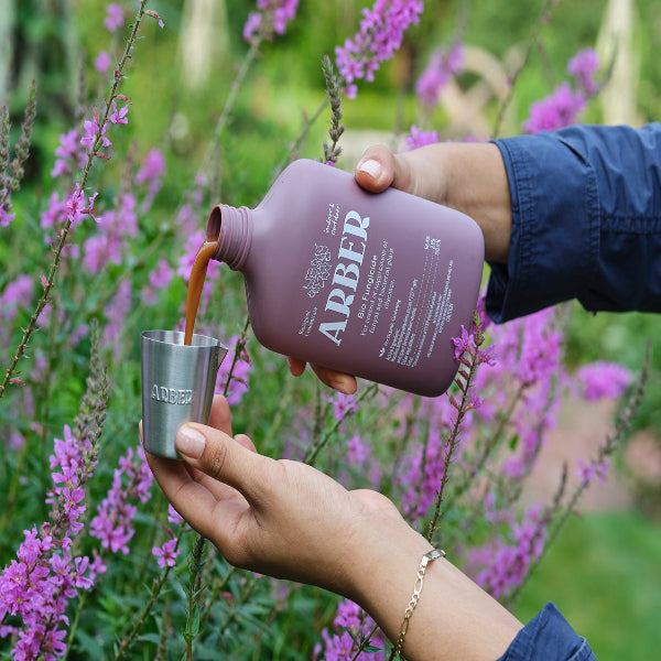 A person pours Arber Organic Bio Fungicide, a brown liquid, from a purple bottle into a metal cup amid blooming purple flowers and lush greenery.