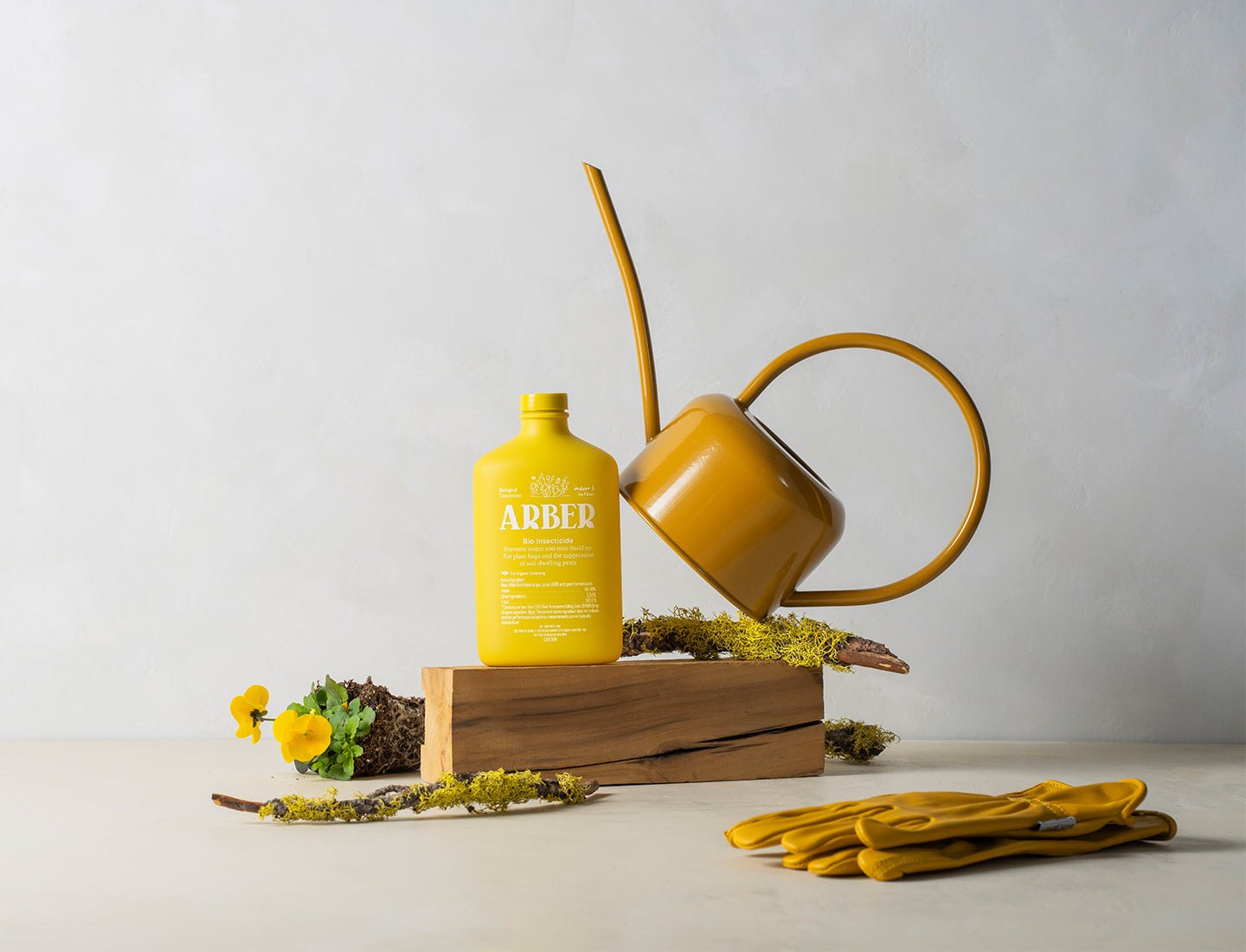 A yellow watering can, a bottle of Arber Organic Bio Insecticide, garden gloves, moss, and small flowers are displayed on a wooden block against a neutral background for natural pest control.