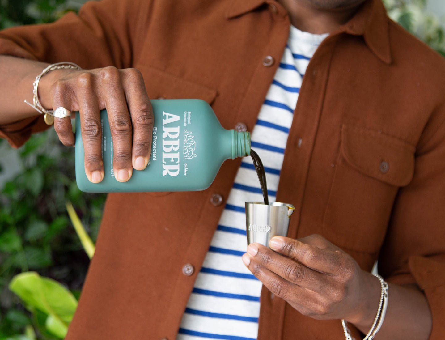 A person in a brown jacket pours Arber Organic Bio Protectant from a green bottle into a small metal cup, with greenery in the background.