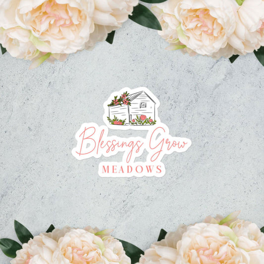 A floral border of pale pink peonies frames the Blessings Grow Meadows logo—a house with flowers and text in pink fonts—on these durable, bubble-free vinyl stickers by Blessings Grow Meadows.