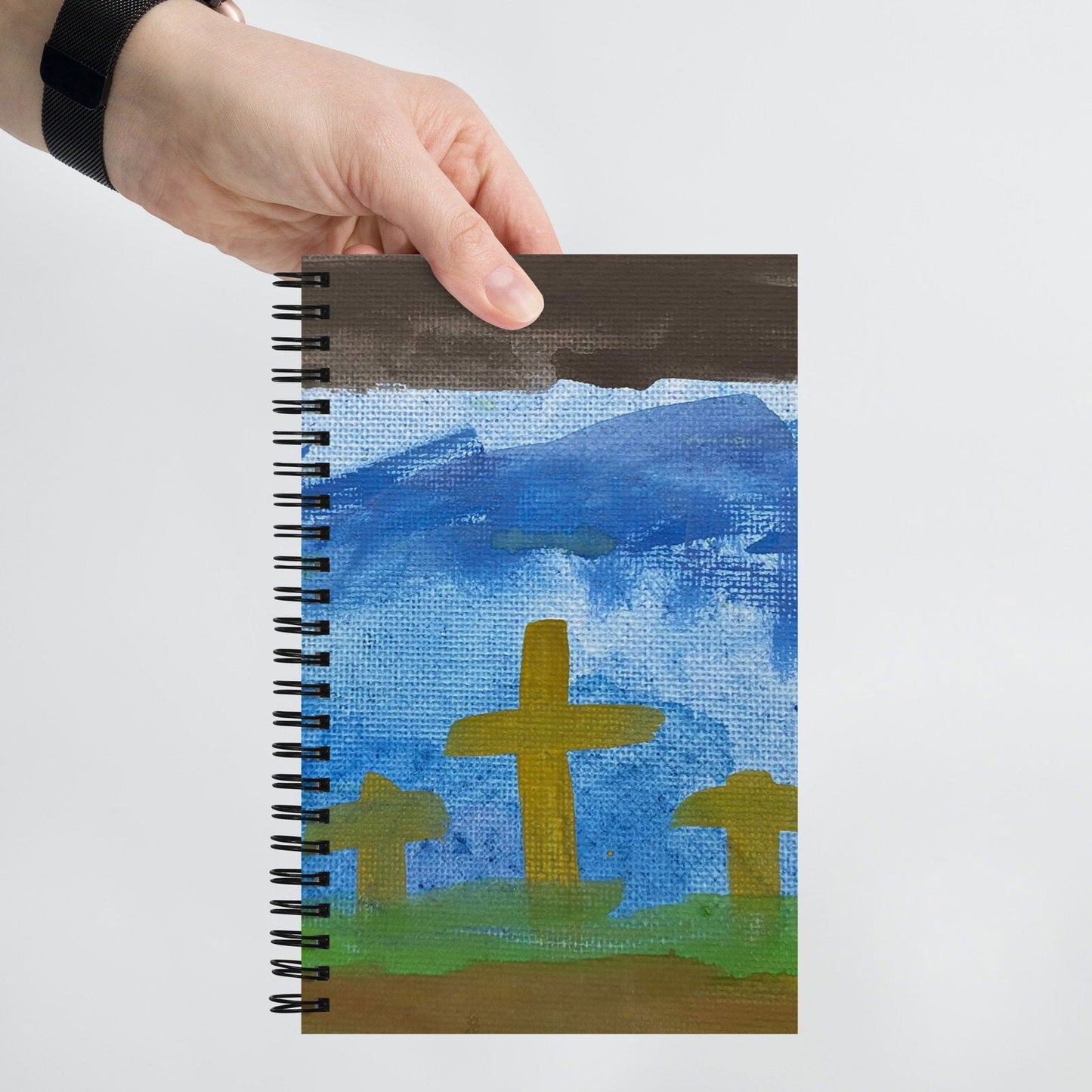 A hand holds the "He is Not Here Original Watercolor Design" spiral notebook by Blessings Grow Meadows, featuring three yellow crosses on green grass beneath a blue sky with dark clouds.