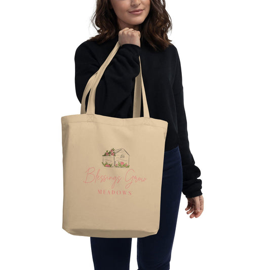 Canvas tote bag featuring Blessings Grow Meadows design with a beige background, pink floral accents, and a charming house illustration.
