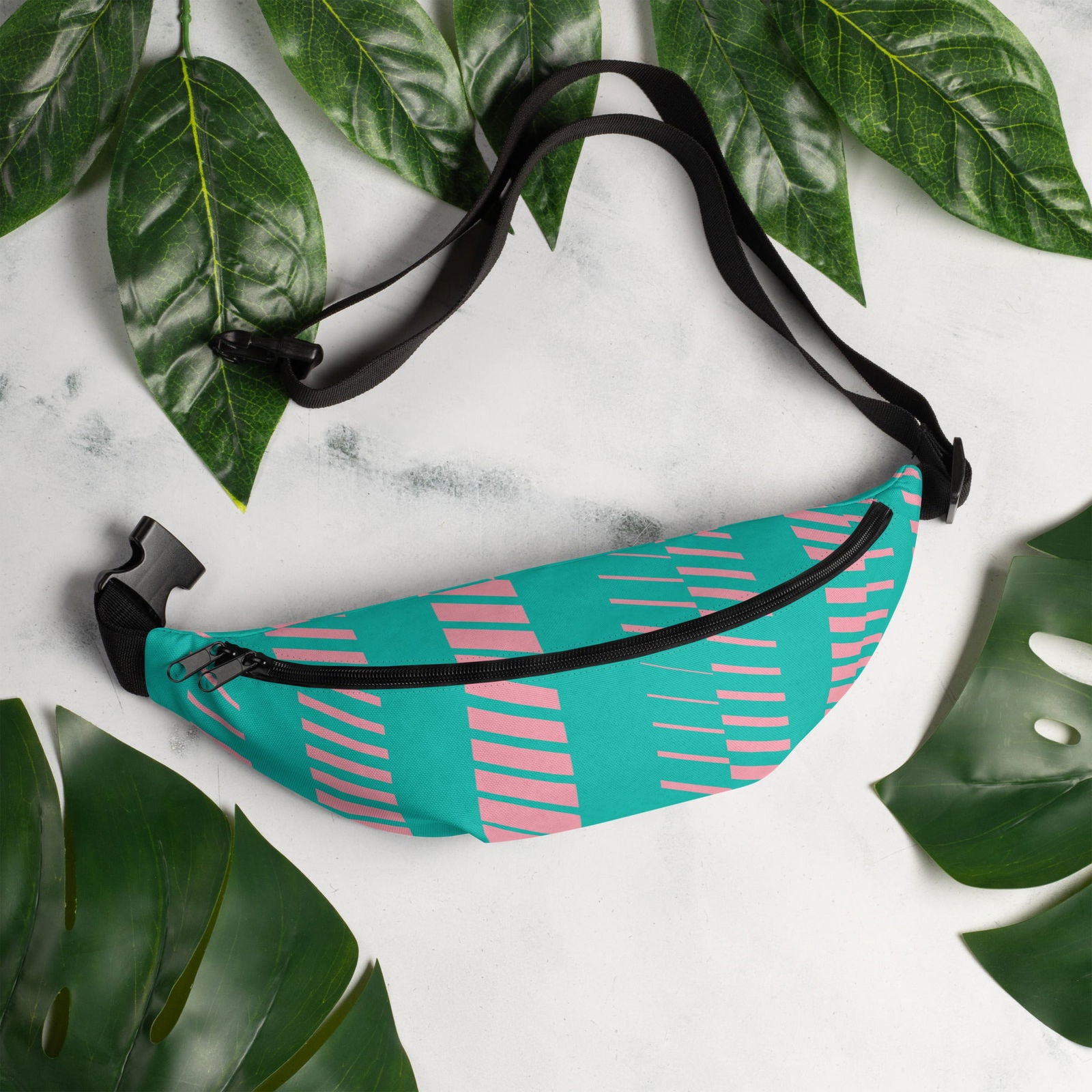 Fabric fanny pack on a white background surrounded by green leaves featuring a teal and pink geometric design with a black zipper and adjustable strap
