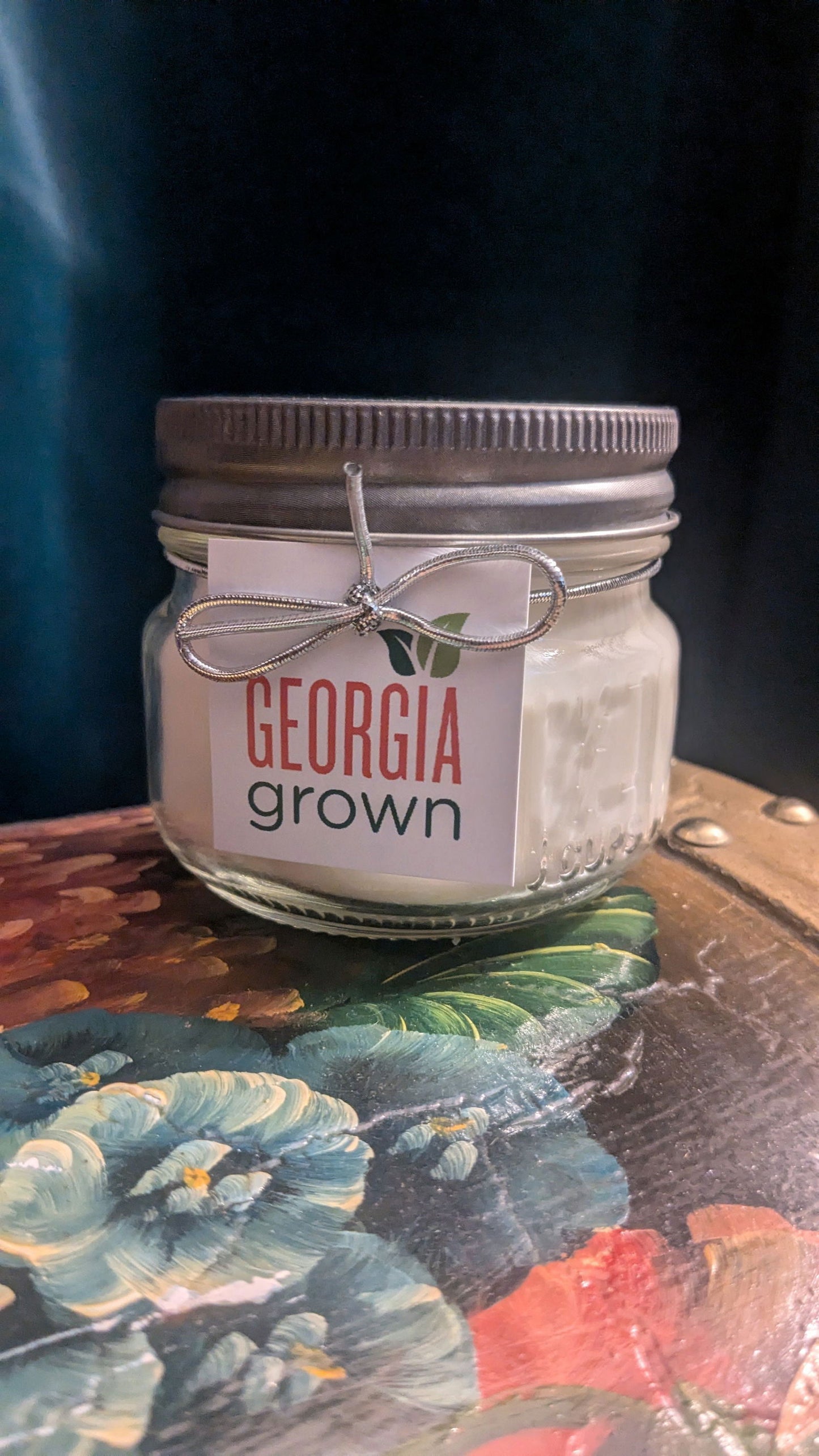 Handmade candle in a mason jar with a silver lid and Georgia Grown label sitting on a vibrant floral painting with teal pansies and red poppies