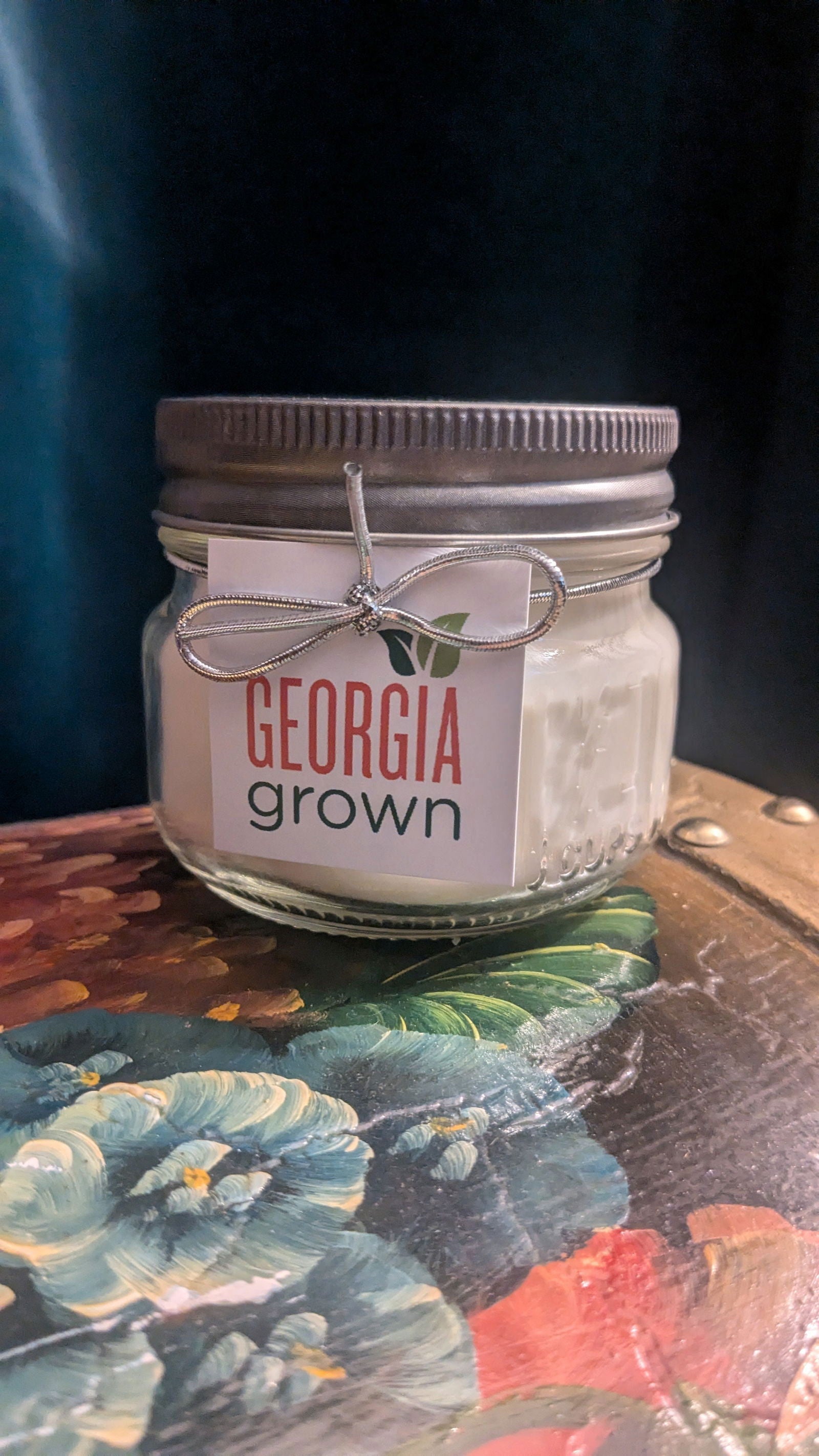 Handmade candle in a mason jar with a silver lid and Georgia Grown label sitting on a vibrant floral painting with teal pansies and red poppies