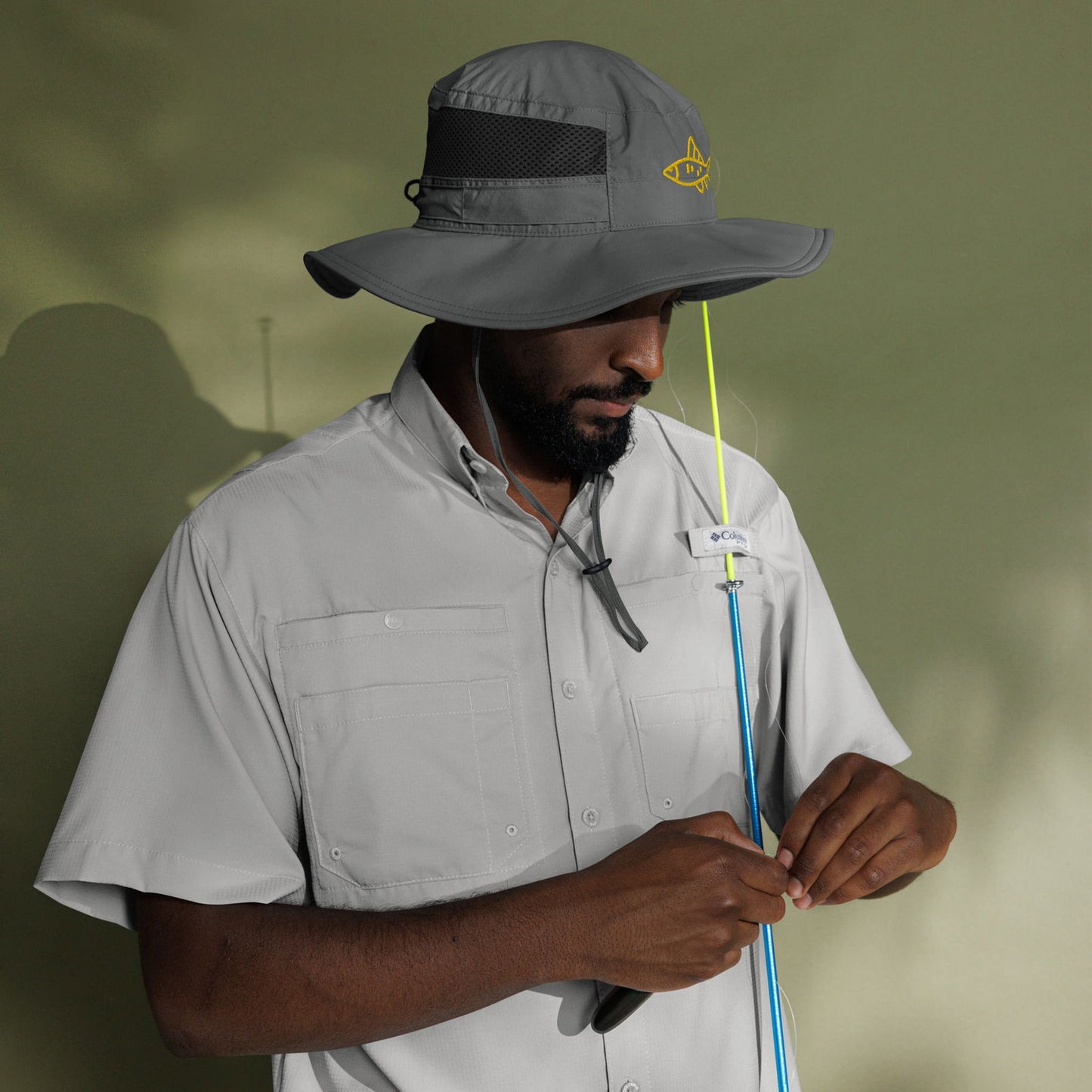 Columbia PFG fishing shirt and hat model preparing fishing rod against a muted green background featuring a bright lime green fishing line and embroidered fish on the hat.