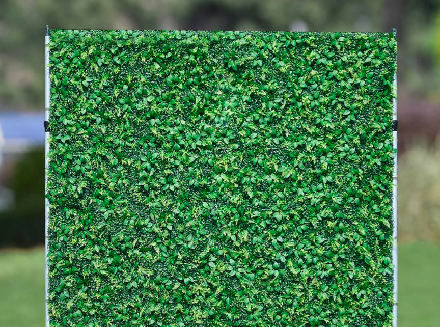 Artificial greenery wall featuring a lush mix of green leaves and subtle textures displayed outdoors against a blurred background of greenery

