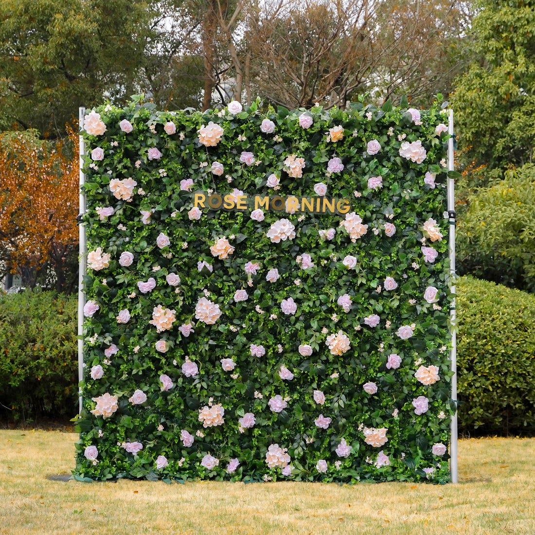 Artificial floral wall installation featuring peach and lavender roses against a backdrop of lush green foliage with the words Rose Morning displayed in gold lettering outdoors near trees

