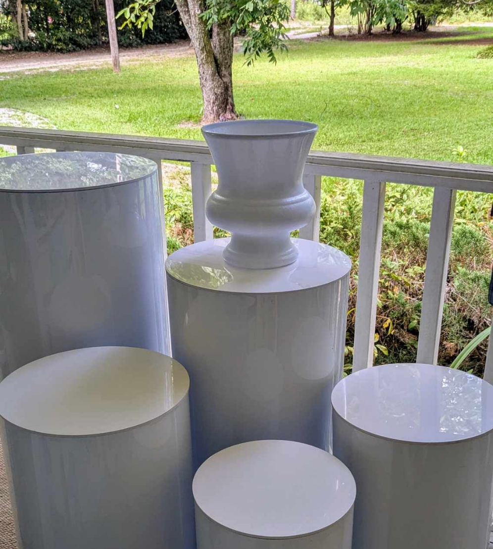 Photograph of four glossy white cylindrical pedestals and a large urn on a porch overlooking a lush green lawn and trees