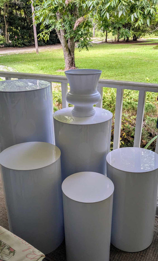 Photograph of five white cylindrical pedestals and an urn on a porch overlooking a lush green lawn and trees
