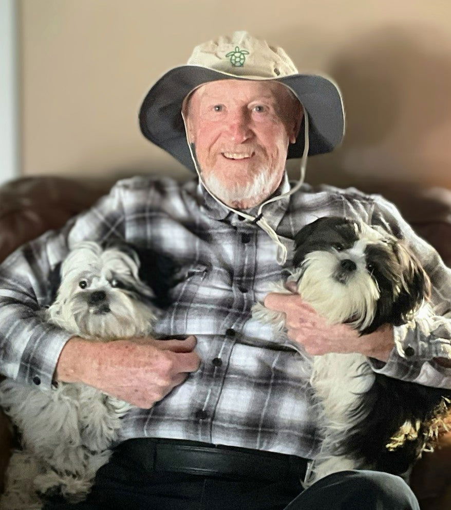 An elderly man wearing a Blessings Grow Meadows Merch Columbia Turtle Boney Hat ~ BGM Custom Designed with UPF 50 sits on a couch, smiling as he hugs two small, fluffy dogs—one white and gray, the other black and white.