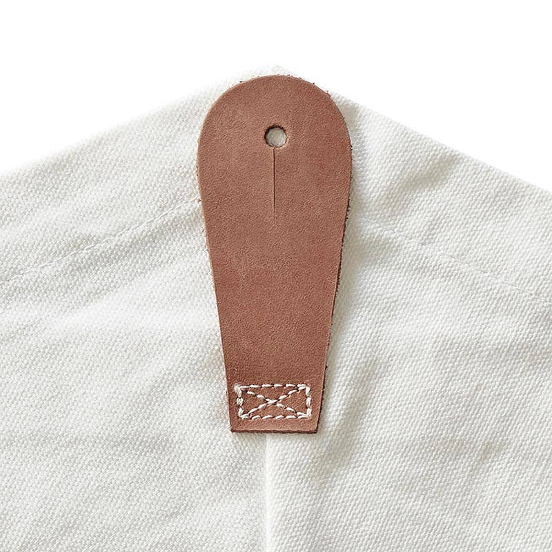 A close-up of a brown leather tab with a hole at the top, stitched onto the Blessings Grow Meadows Merch & More Bouquet Bag - Mama, crafted from white canvas and featuring a floral design.