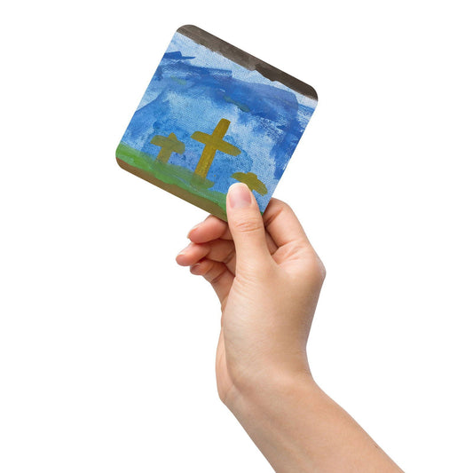 A hand holds the “Cork-back coaster Original Watercolor Cross” by Blessings Grow Meadows Merch & More, showing three crosses on a green hill under a blue sky. The background is plain white, highlighting its religious or Easter theme.