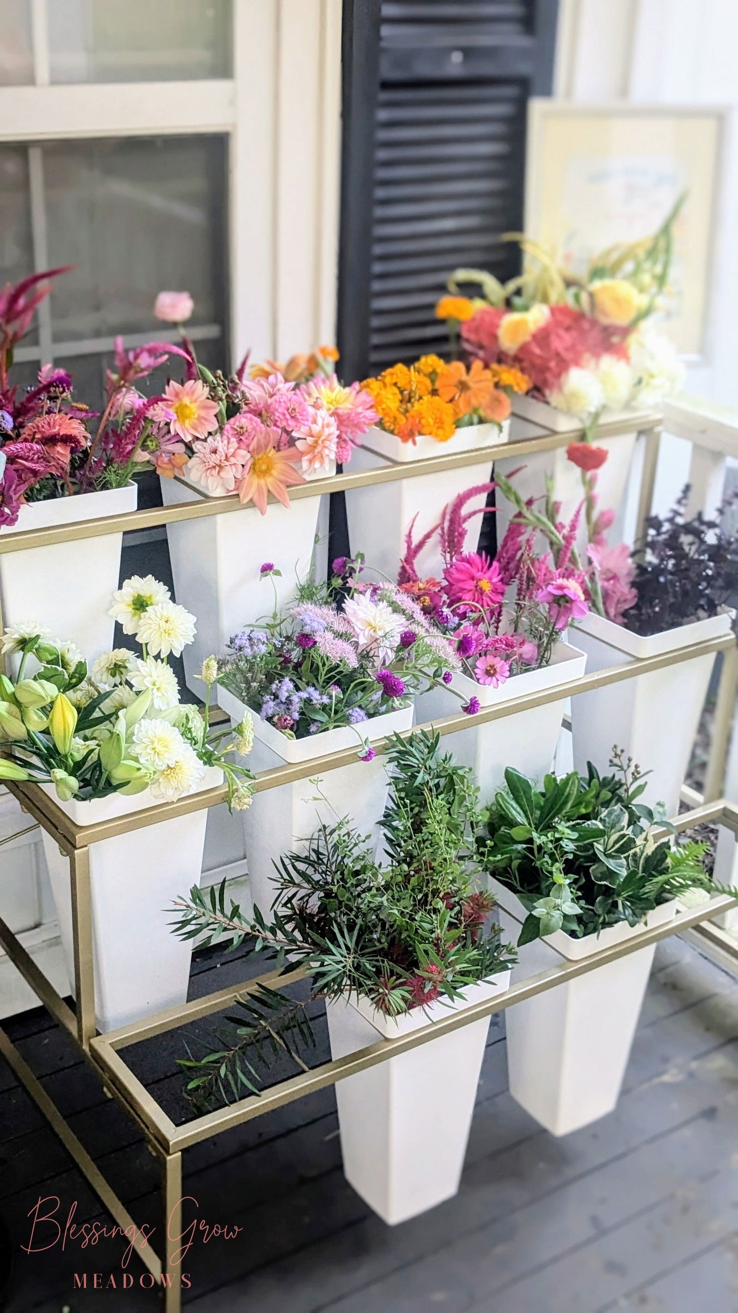 Blessings Grow Meadows photography shows a tiered gold stand holding white vases of colorful dahlias, lilies, and assorted greenery near a window and black shutters
