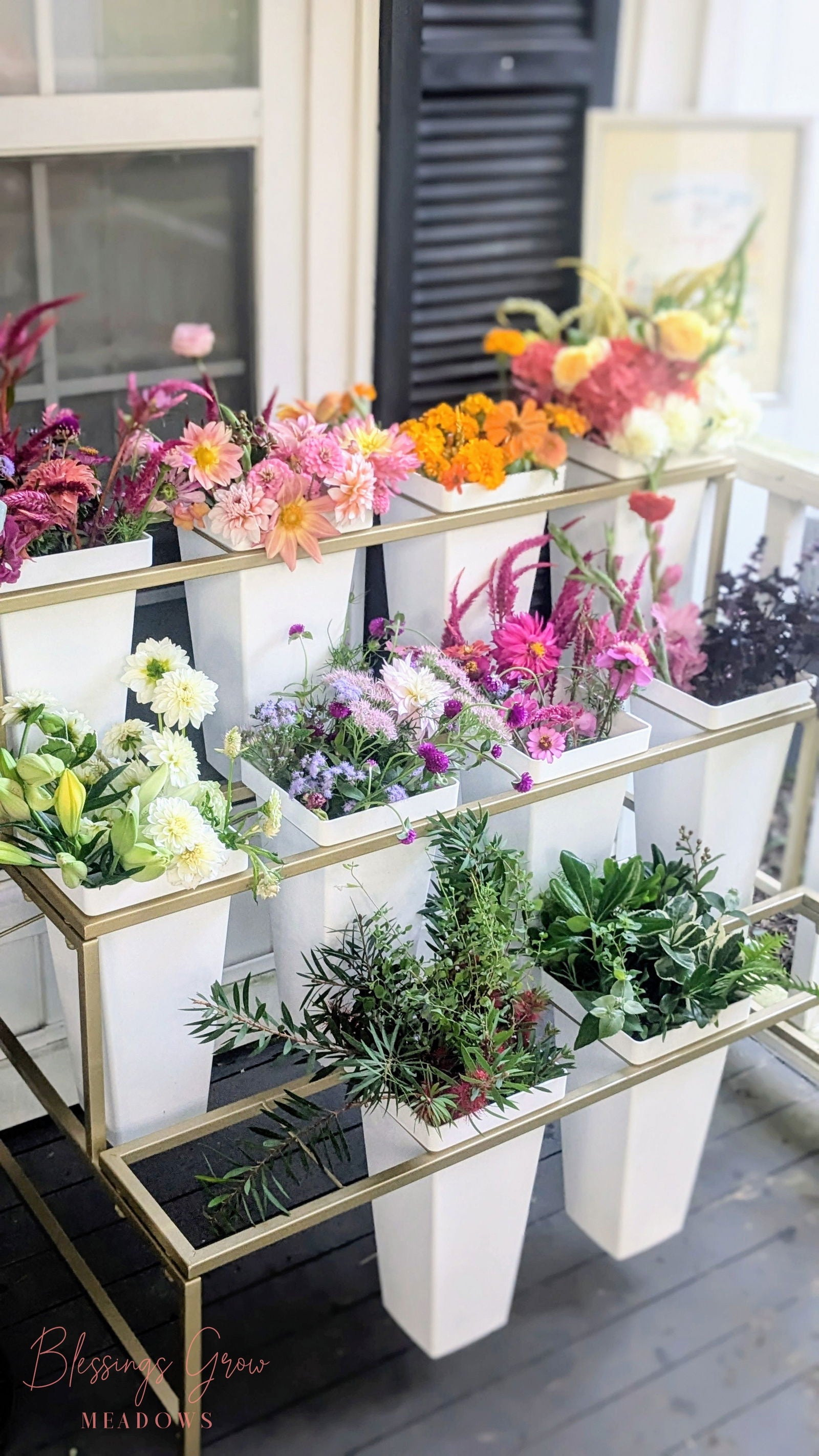 Blessings Grow Meadows photography shows a tiered gold stand holding white vases of colorful dahlias, lilies, and assorted greenery near a window and black shutters
