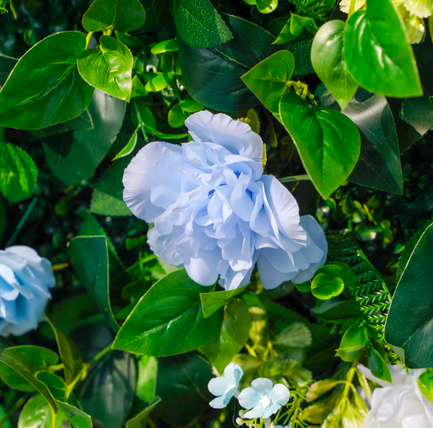 Artificial flower arrangement featuring a large light blue peony amongst lush green leaves and smaller light blue flowers.

