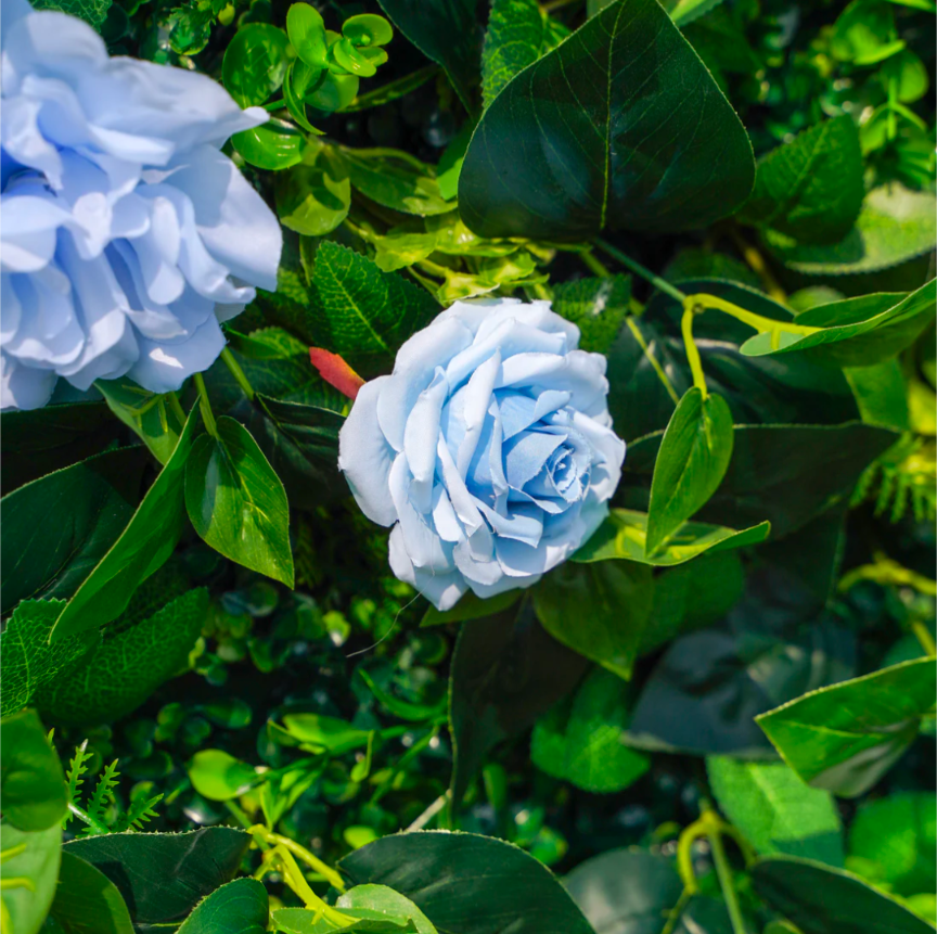 Artificial floral arrangement featuring pastel blue roses and lush green leaves with hints of red stems and varied leaf textures

