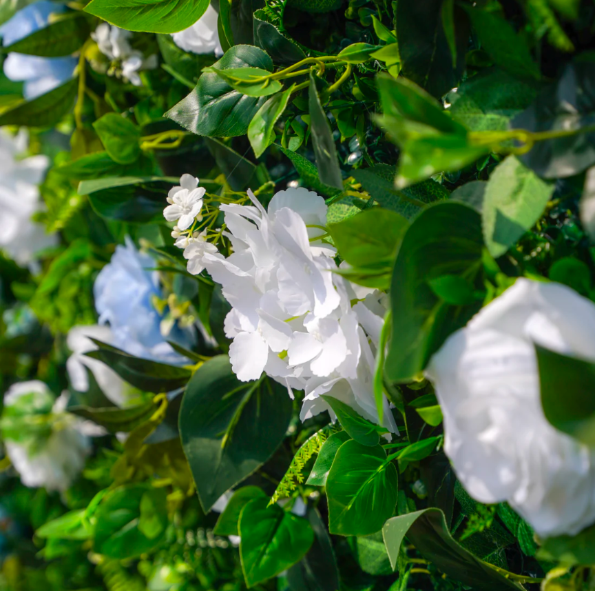 Artificial flower arrangement featuring vibrant white hydrangeas and light blue roses with lush green leaves showcasing a textured floral wall.
