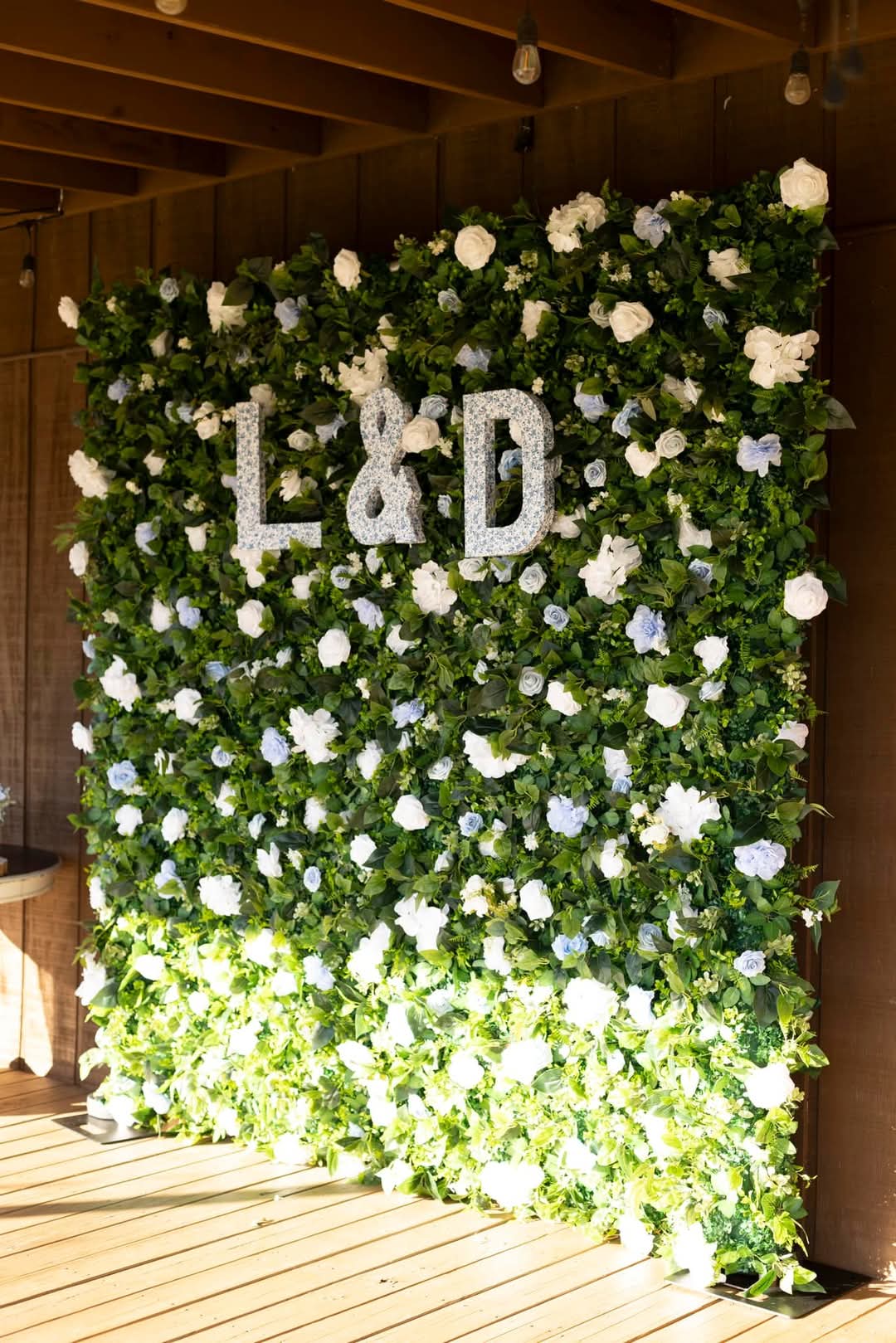 Artificial floral backdrop featuring L&D initials, white and light blue roses, and lush greenery against a rustic wooden wall
