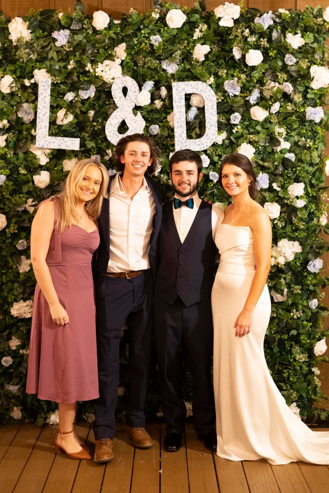 Photograph of four people posing in front of a floral wall featuring the letters L & D mauve dress brown shoes white dress and a teal bow tie
